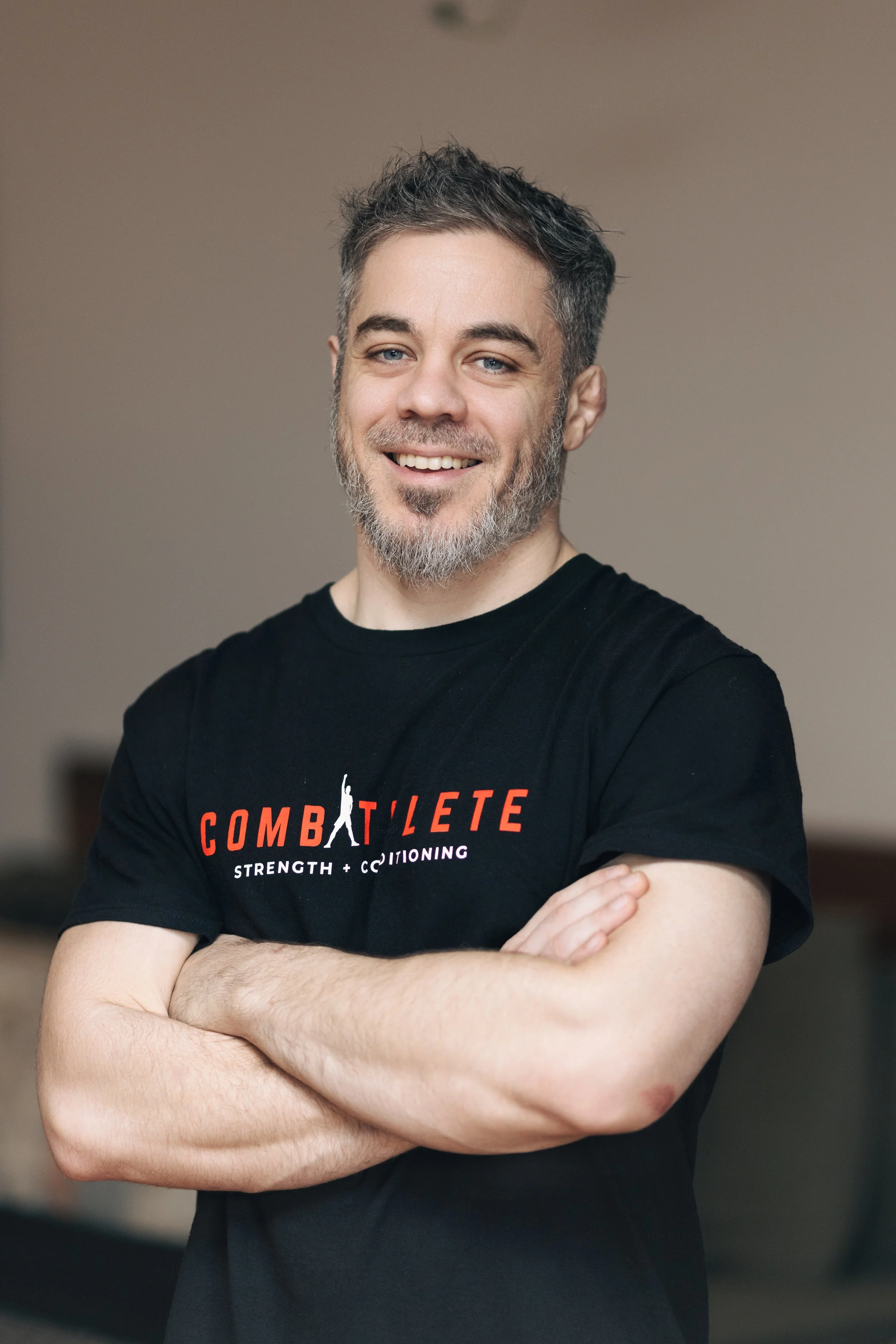 A man with gray hair and beard smiling, arms crossed, wearing a black t-shirt with red and white text for a fitness or strength training brand.