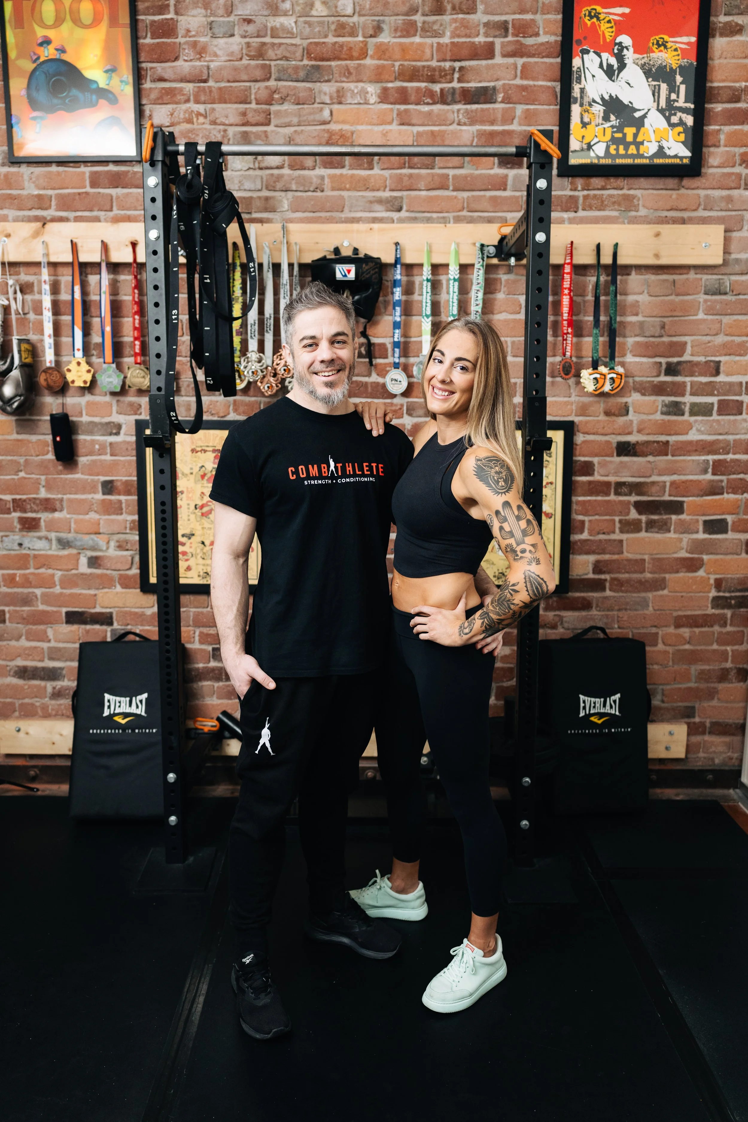 Two smiling fitness enthusiasts, a man and a woman, standing together in a gym with a brick wall background. The man wears a black T-shirt with 'COMBAT ATHLETE' printed on it and black pants. The woman has long blonde hair, a tattoo sleeve on her left arm, and is dressed in a black sports bra and matching leggings. They stand close with their arms around each other's shoulders, in front of a power rack with medals and posters on the wall.