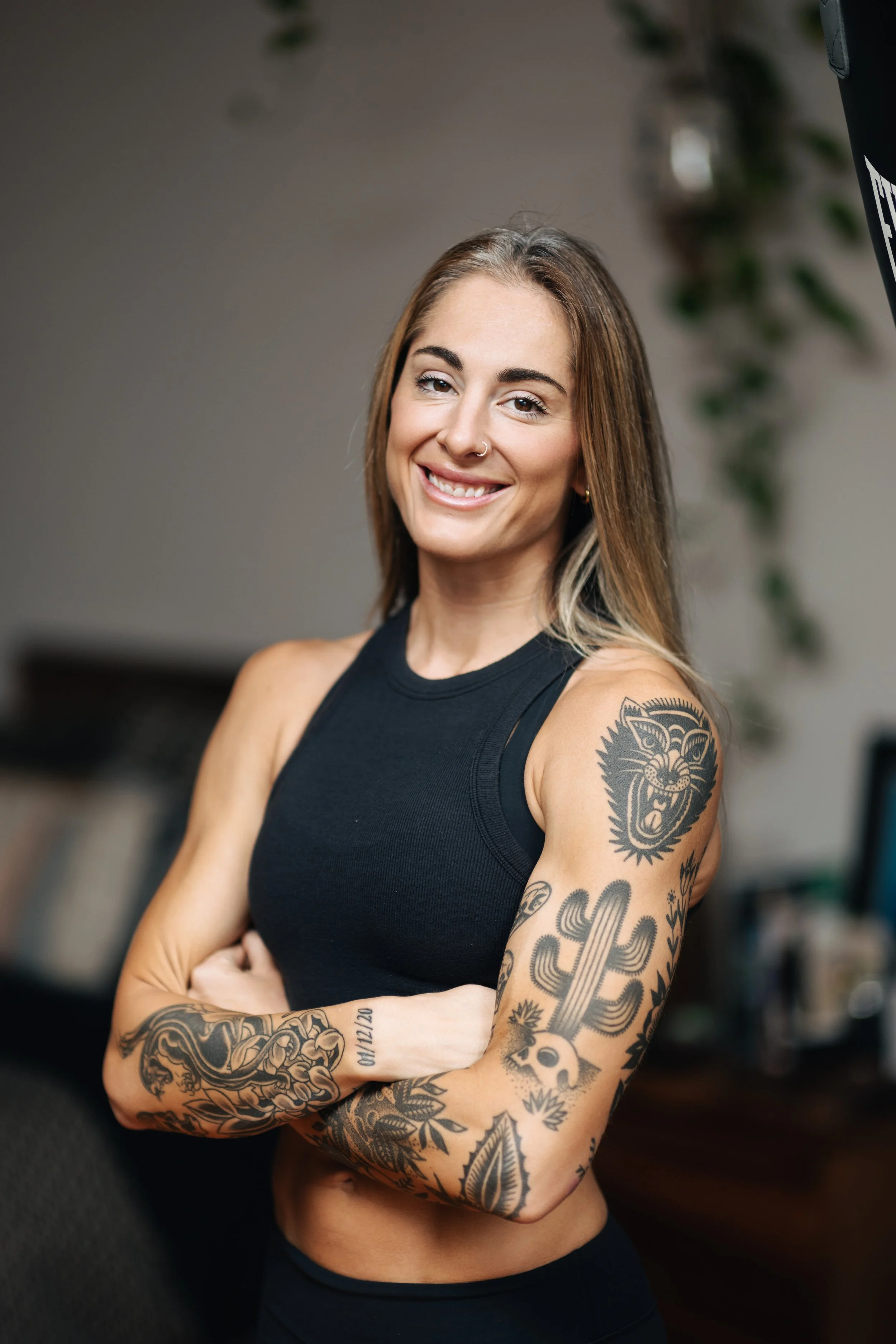 A smiling woman with tattoos on her arms and chest, wearing a black sleeveless top, standing with her arms crossed indoors with a blurred background.