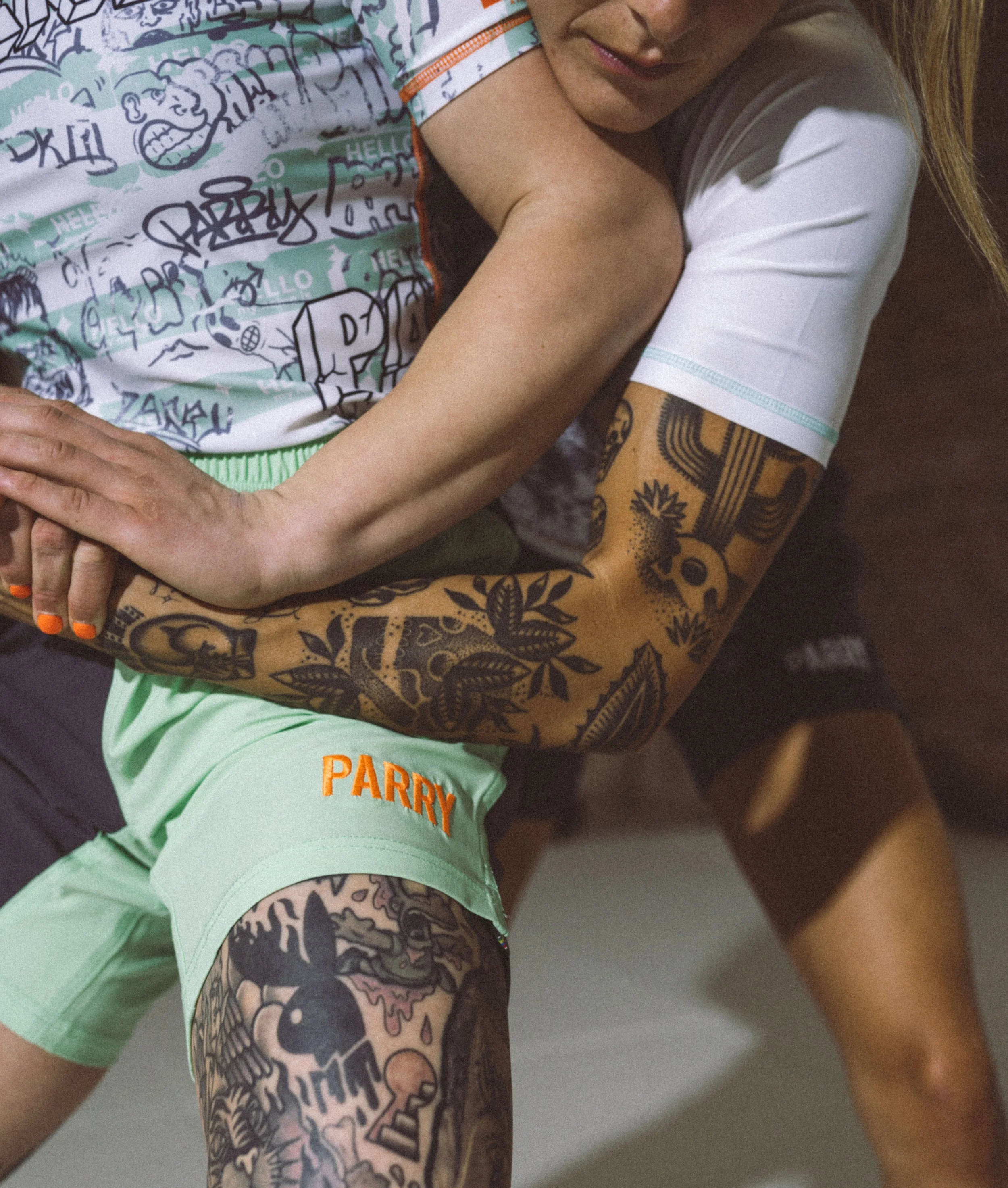 Close-up of a person with tattoos on their arm and tattoos on their thigh, wearing light green shorts and a white shirt, hugging a child wearing a colorful t-shirt with cartoon-style drawings and text, smiling softly.