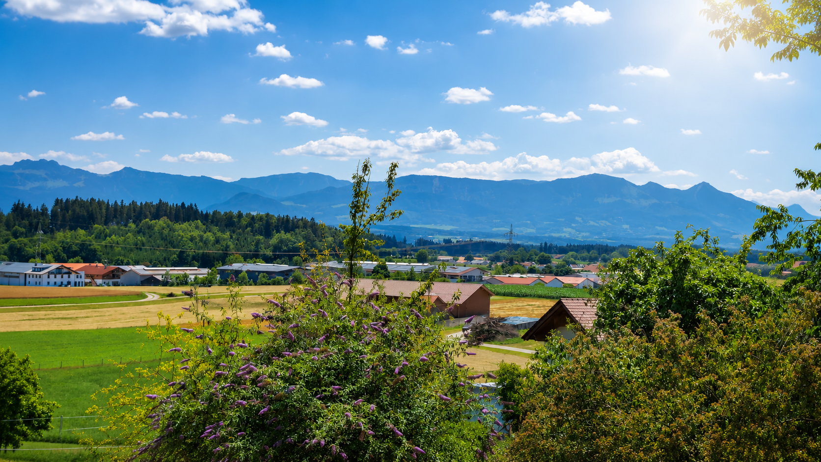 Blick auf eine grüne Landschaft mit Feldern, Bäumen und Häusern vor einer Bergkette bei sonnigem Wetter.