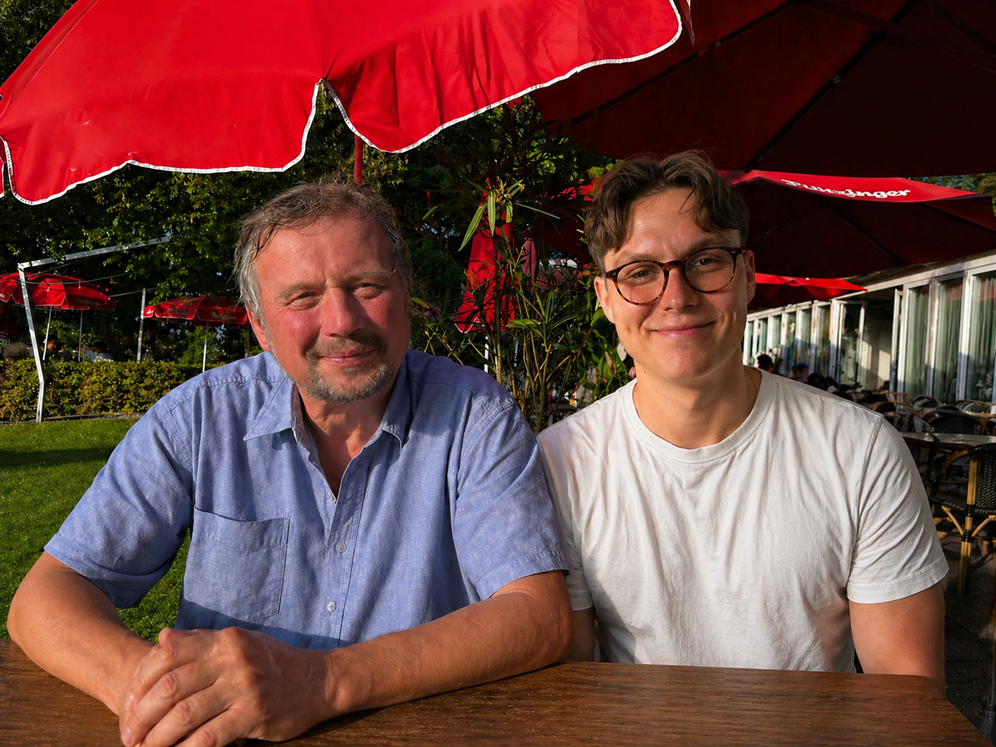 Zwei Männer sitzen an einem Tisch im Außenbereich eines Cafés unter roten Sonnenschirmen, lachend und entspannt, im Sonnenlicht, mit Büschen im Hintergrund.