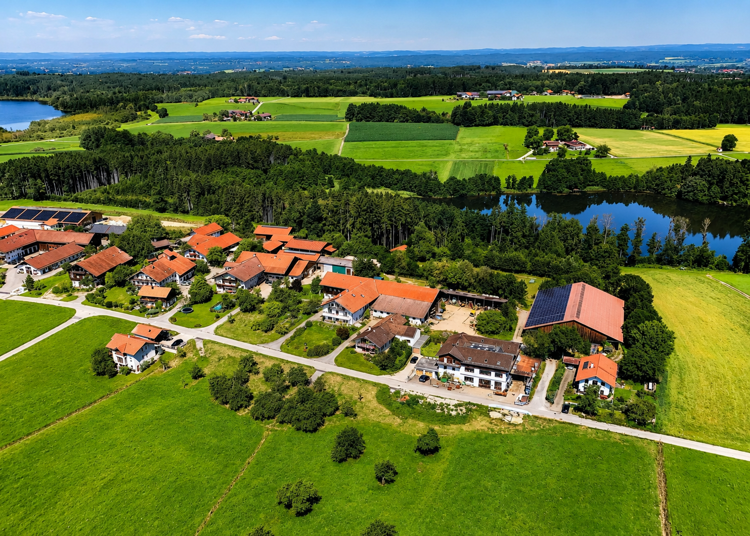 Luftaufnahme eines Dorfes mit Häusern, umgeben von Wiesen, Wäldern, Seen und Feldern in einer ländlichen Gegend.