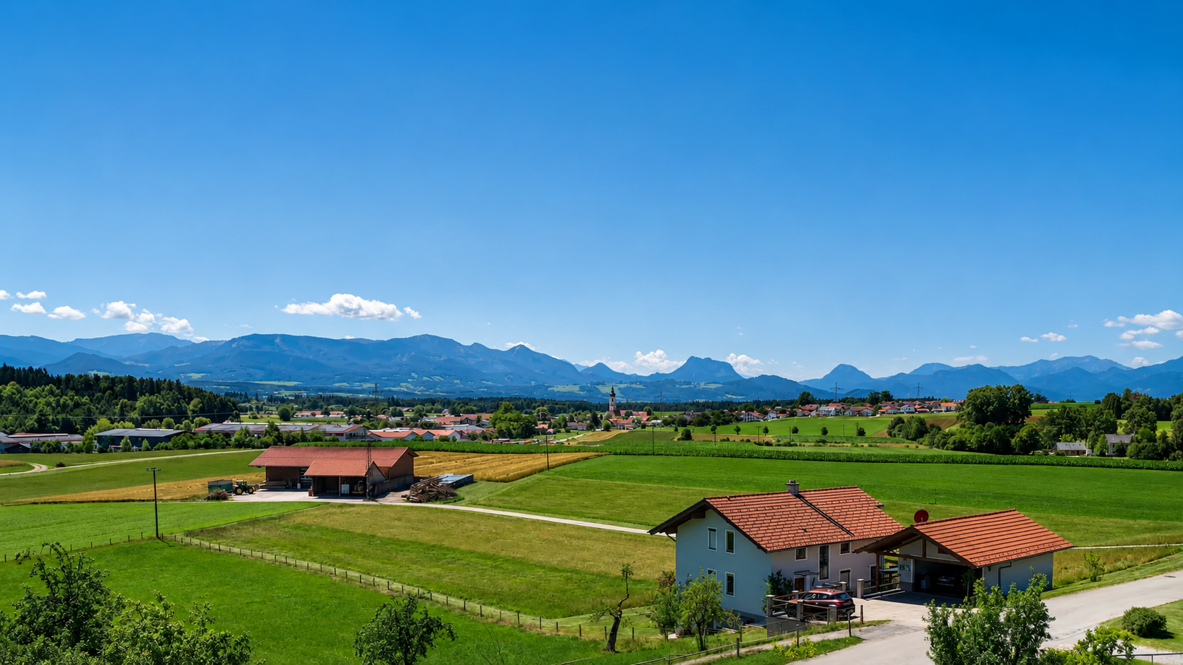 Landschaft mit grünem Feld, Häusern und Bergen im Hintergrund unter blauem Himmel mit wenigen Wolken.