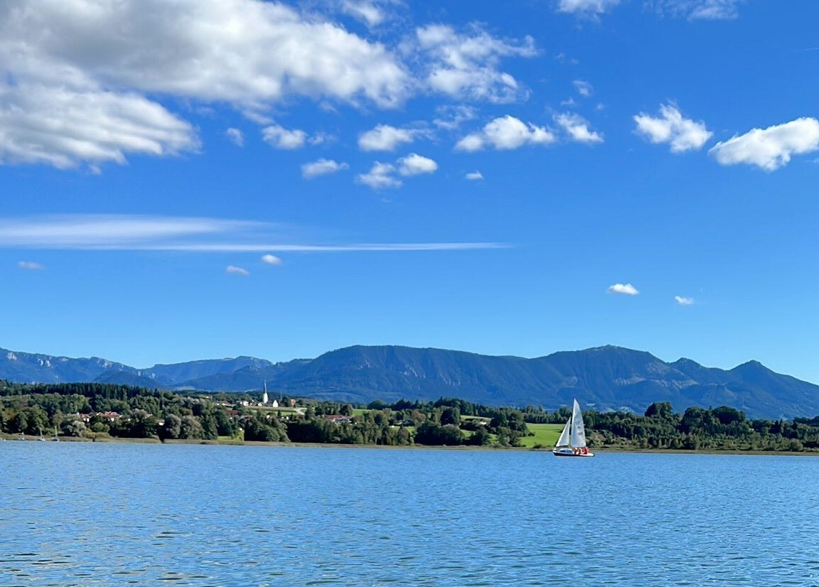 Blick auf einen See mit Segelboot, grüne Bäume am Ufer und Berge im Hintergrund unter einem blauen Himmel mit Wolken.