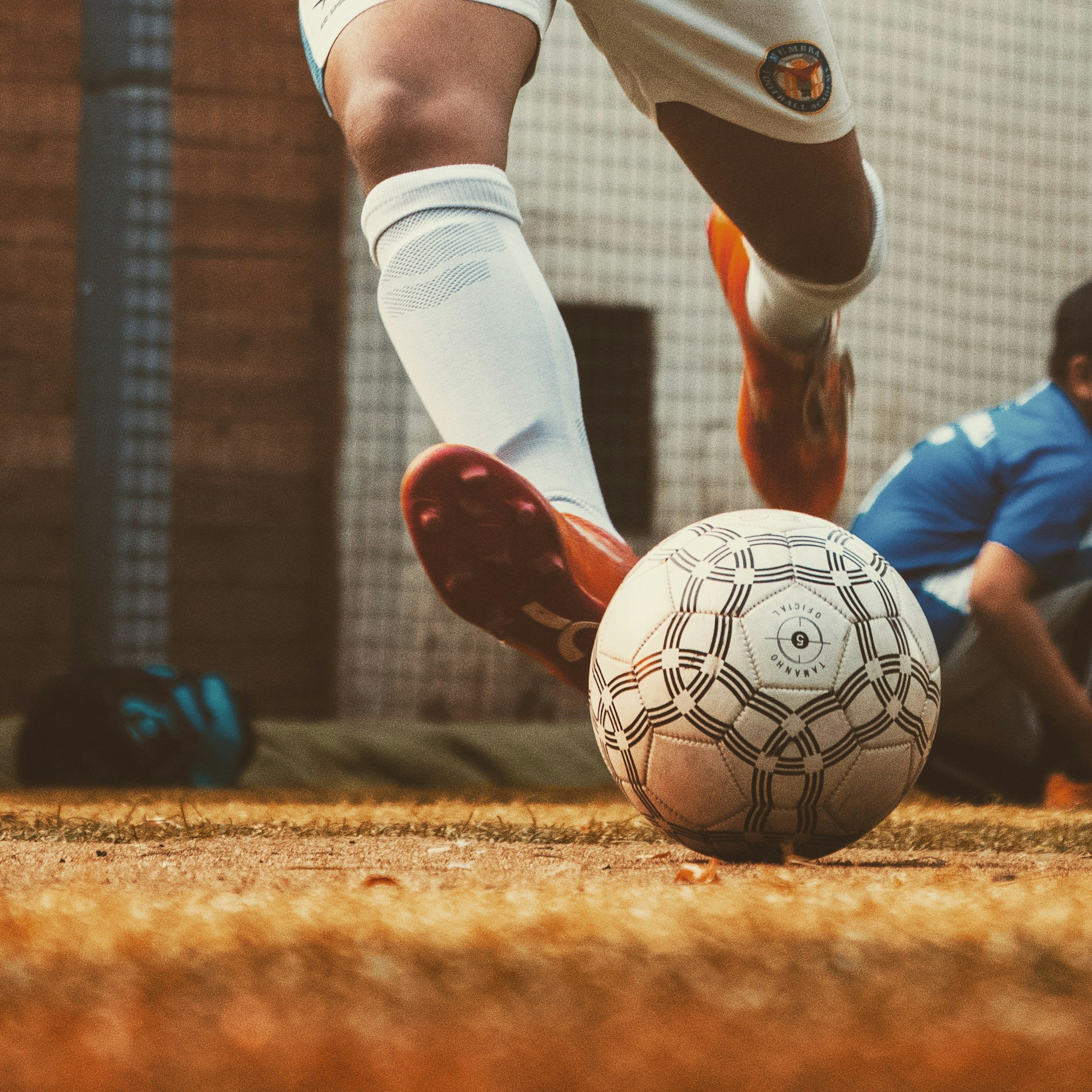 Fußball liegt auf einem warmen Erdboden, im Hintergrund sind die Füße eines Spielers in Sportnoten zu sehen, der in einem weißen Trikot mit blauem Akzent steht.