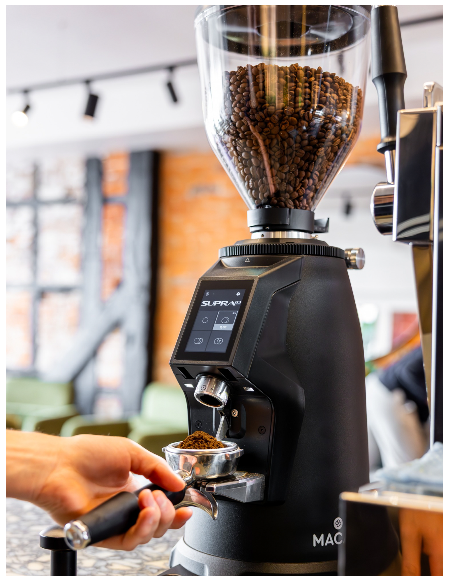 Person tamping ground coffee into a portafilter beneath a coffee grinder filled with roasted coffee beans in a café setting.