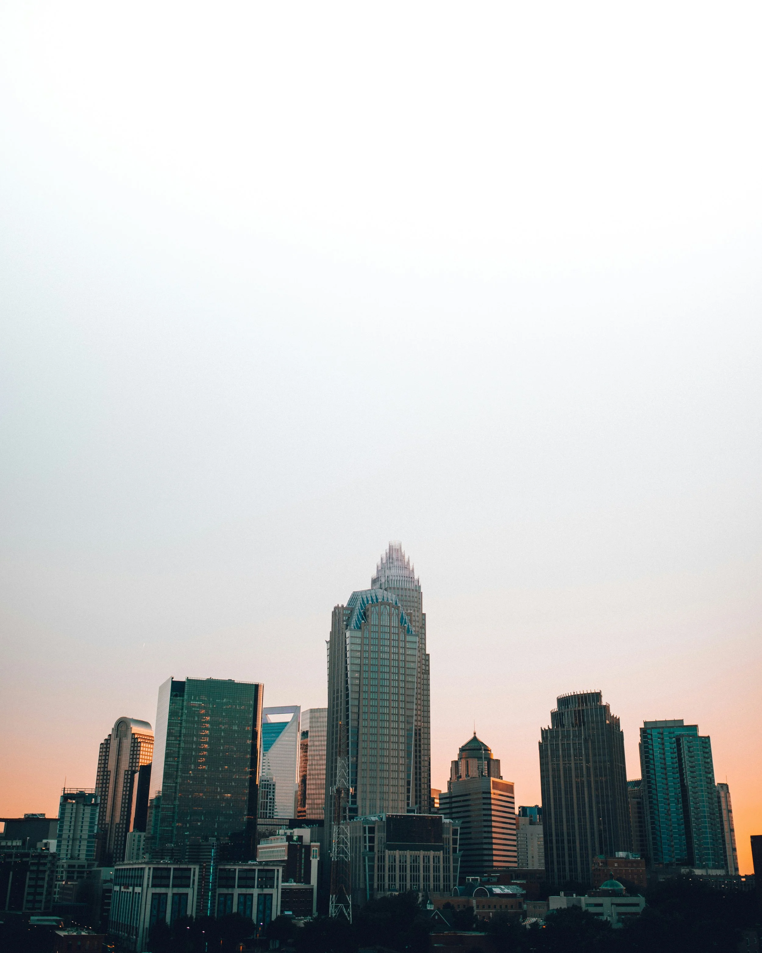 Skyline of a city with skyscrapers at sunset, featuring a tall building with a pointed top and blue accents, surrounded by other high-rise buildings.