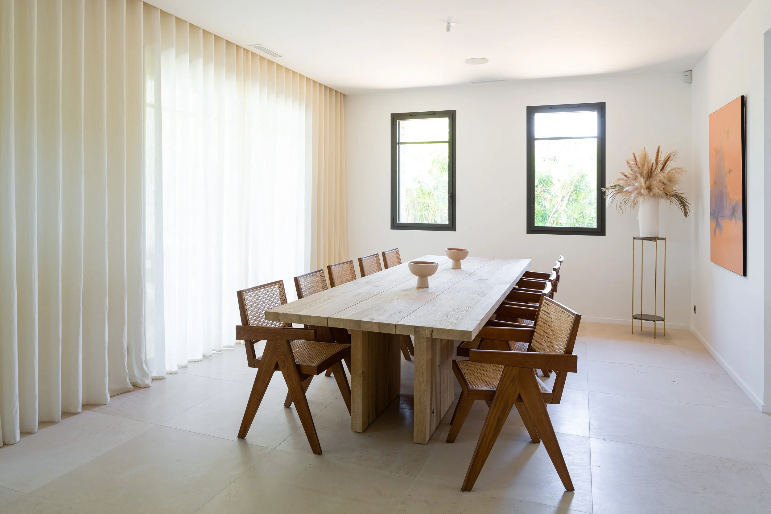 Salle à manger moderne avec une grande table en bois et des chaises en bois. Deux fenêtres lumineuses, un mur blanc, un arbre décoratif avec des plantes séchées dans un vase blanc, et un tableau orange sur le mur.