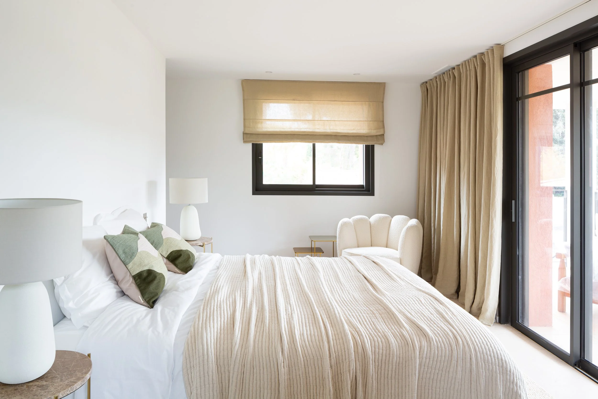 Chambre moderne avec lit, lampe de chevet, et grandes fenêtres avec rideaux beige, donnant sur un balcon.