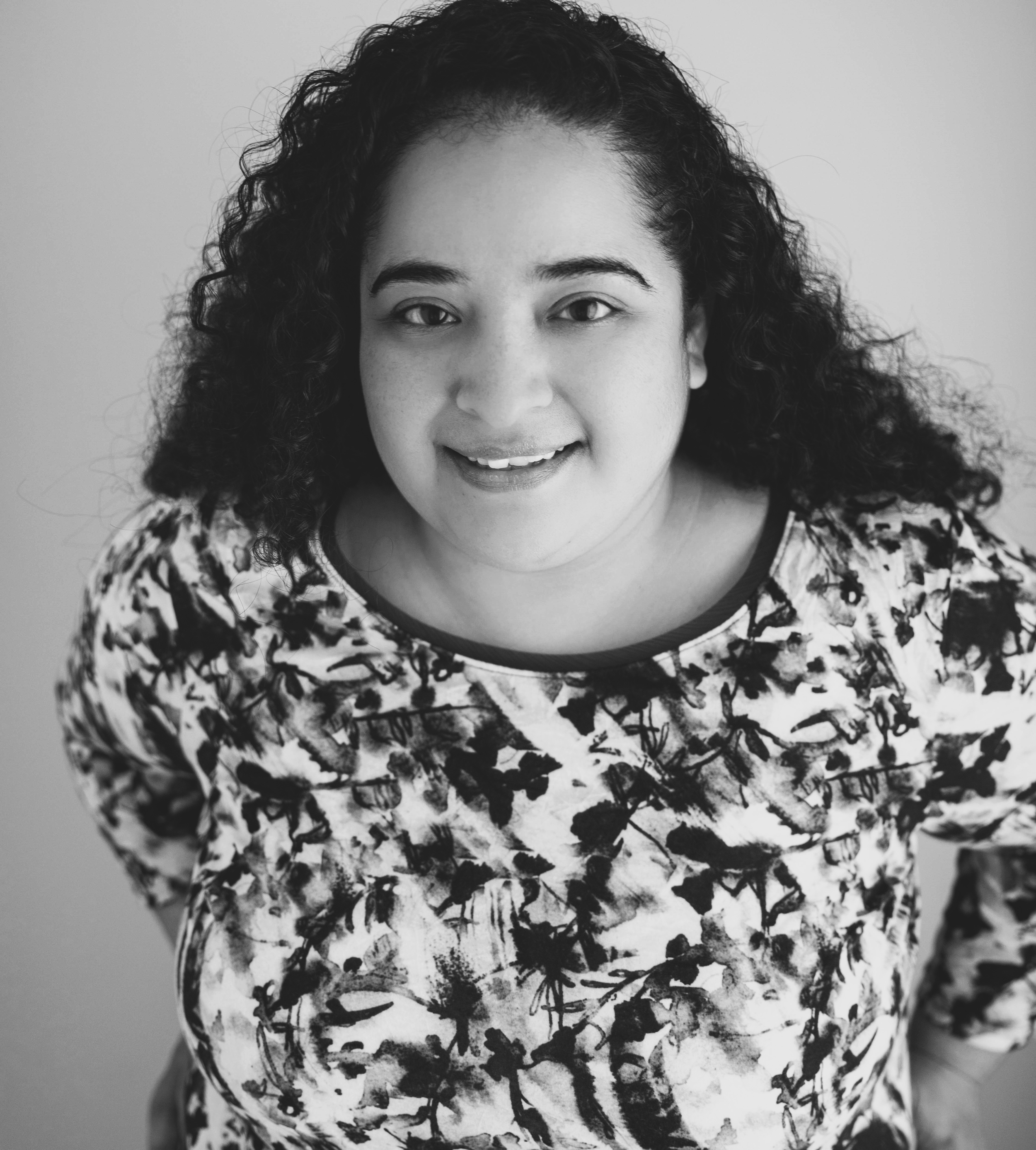 Black and white portrait of a young woman with curly hair smiling, wearing a printed top with a black neckline.