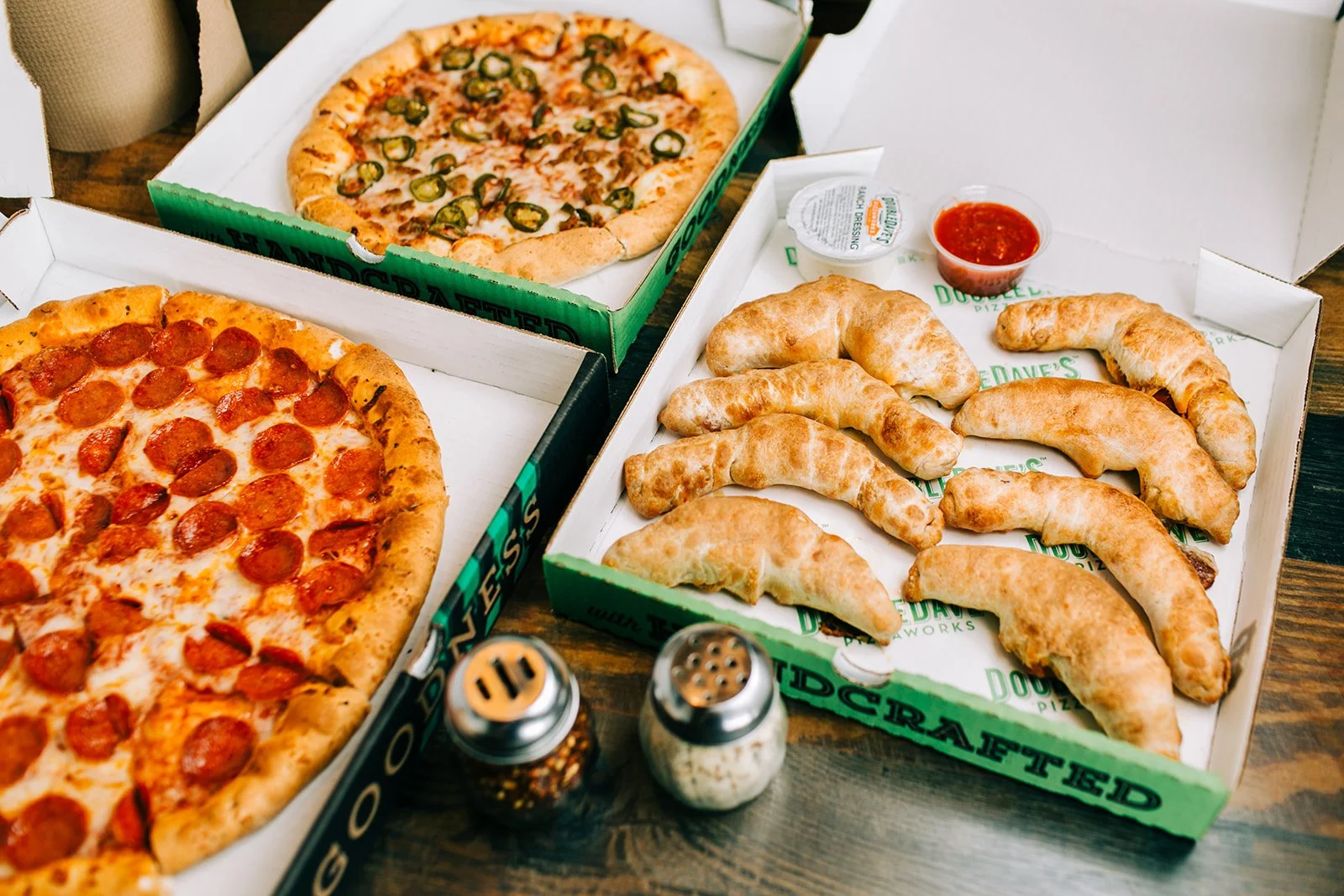 Two pizza boxes with pepperoni and jalapeño pizzas, a box of fried croissants with a side of marinara sauce, salt and pepper shakers, on a wooden table.