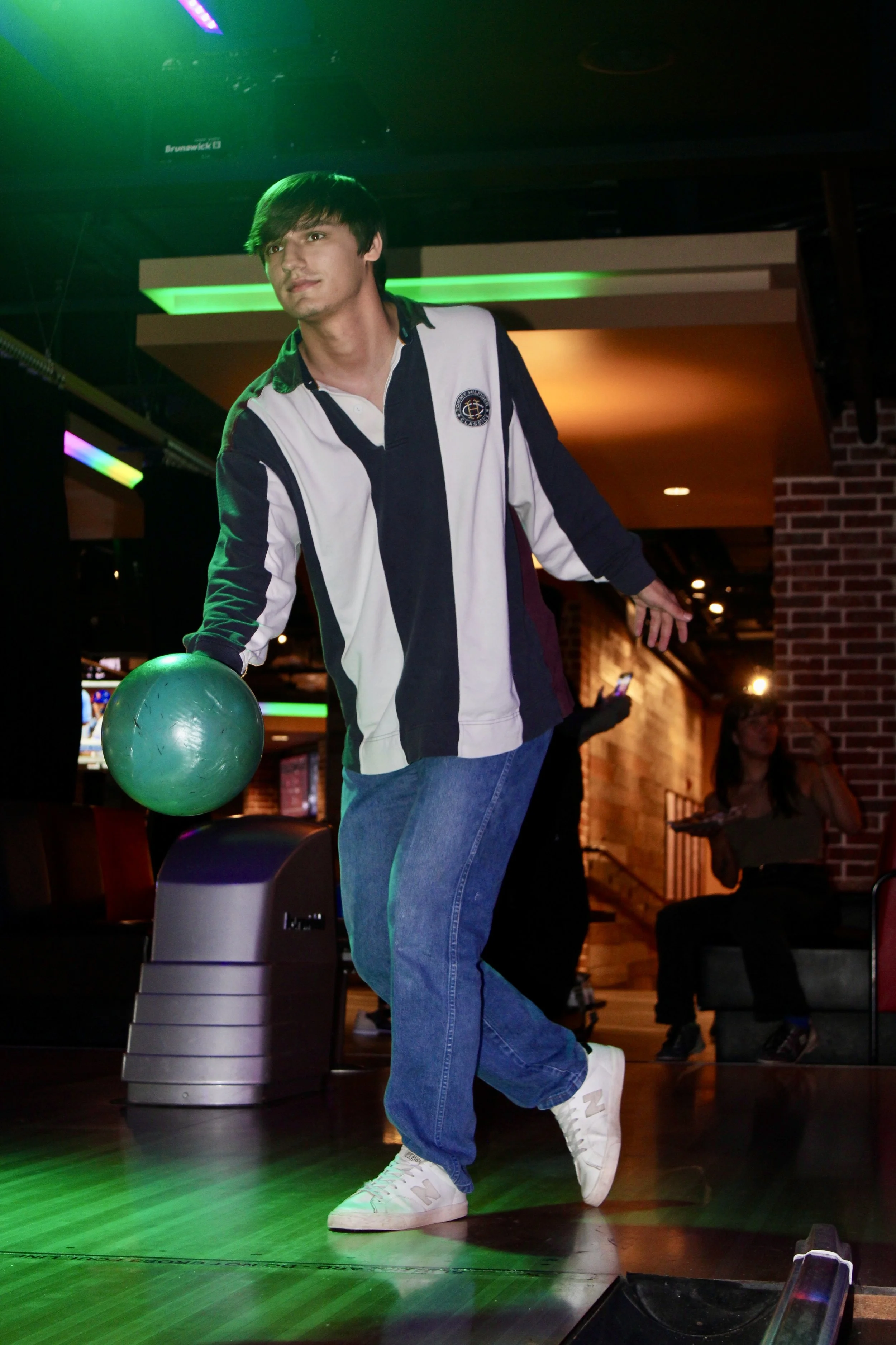 A young man extending his arm with a bowling ball in his hand at a bowling alley, with a woman sitting in the background drinking from a glass.