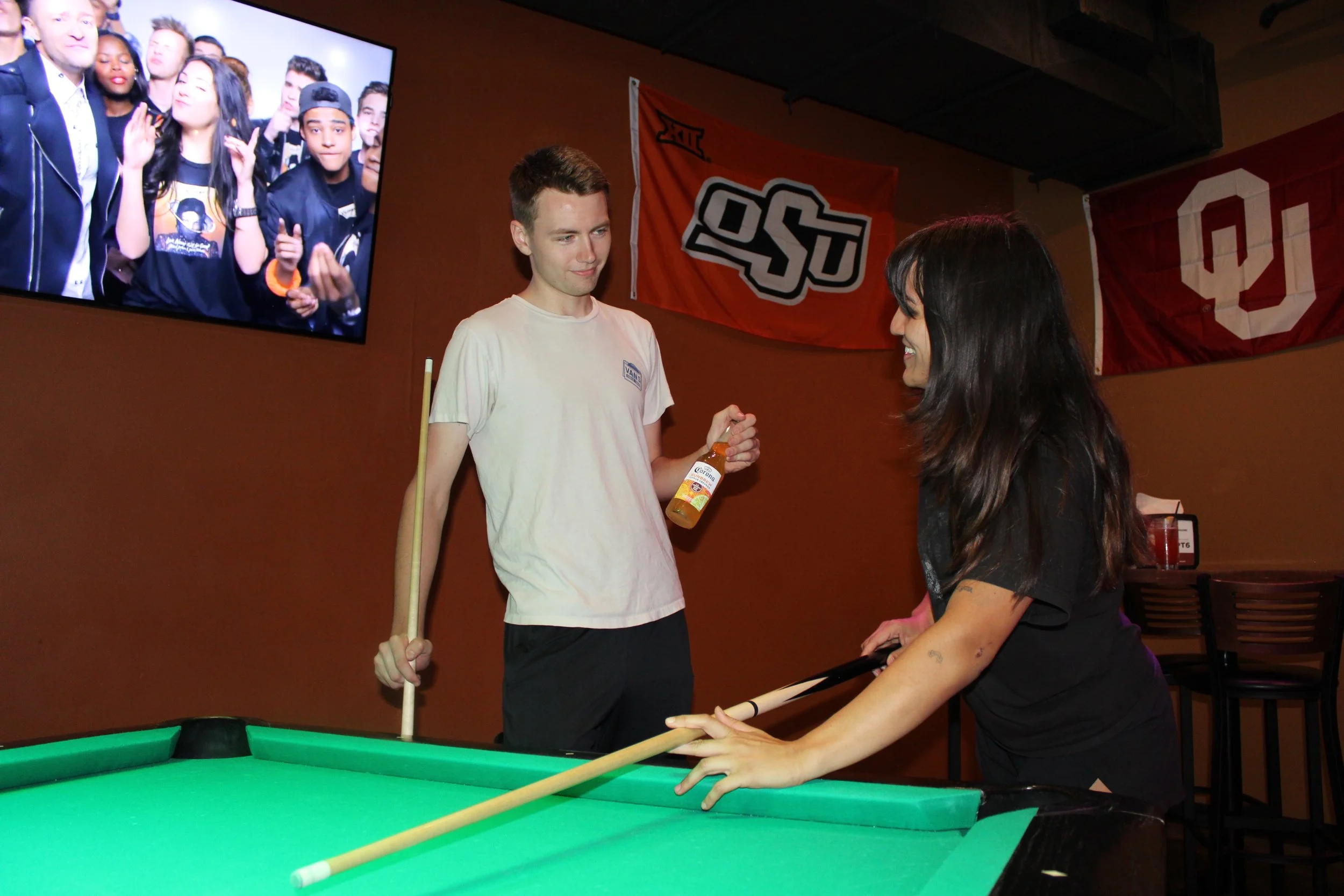 A young woman and a young man play pool at a bar. The woman is smiling and leaning on the pool table, holding a pool cue, while the man is holding a beer and a pool cue, looking at her. There are Oklahoma and Big 12 banners hanging on the wall, and a TV screen showing a group of people in the background.