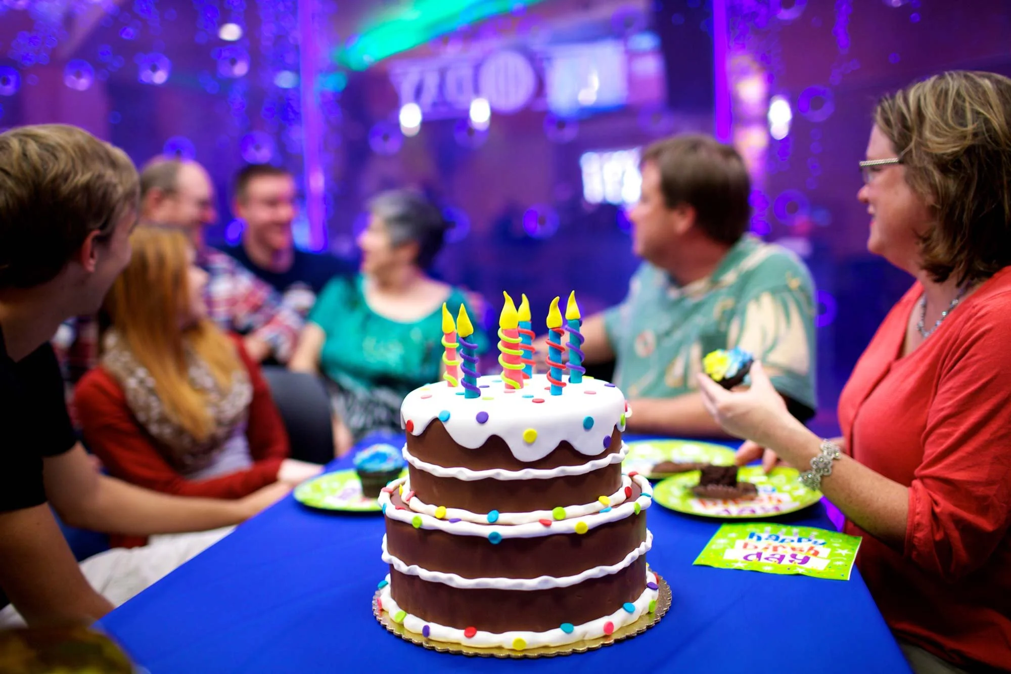 A birthday party with a chocolate cake decorated with white icing, colorful sprinkles, and lit birthday candles, surrounded by people celebrating indoors with purple and blue lighting.