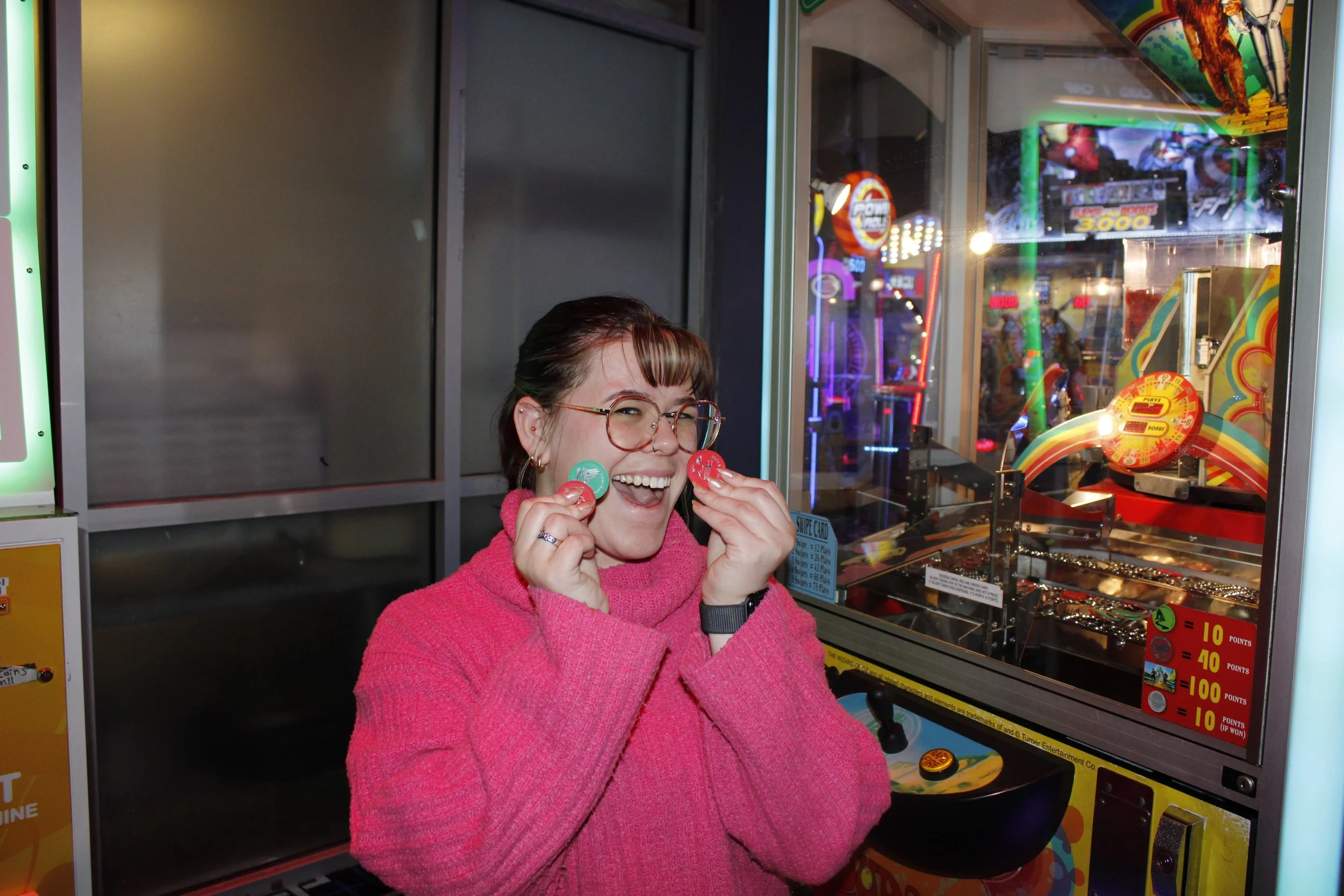 A young woman with glasses and a pink sweater smiling and holding red and green tokens in front of an arcade claw machine filled with toys and prizes.