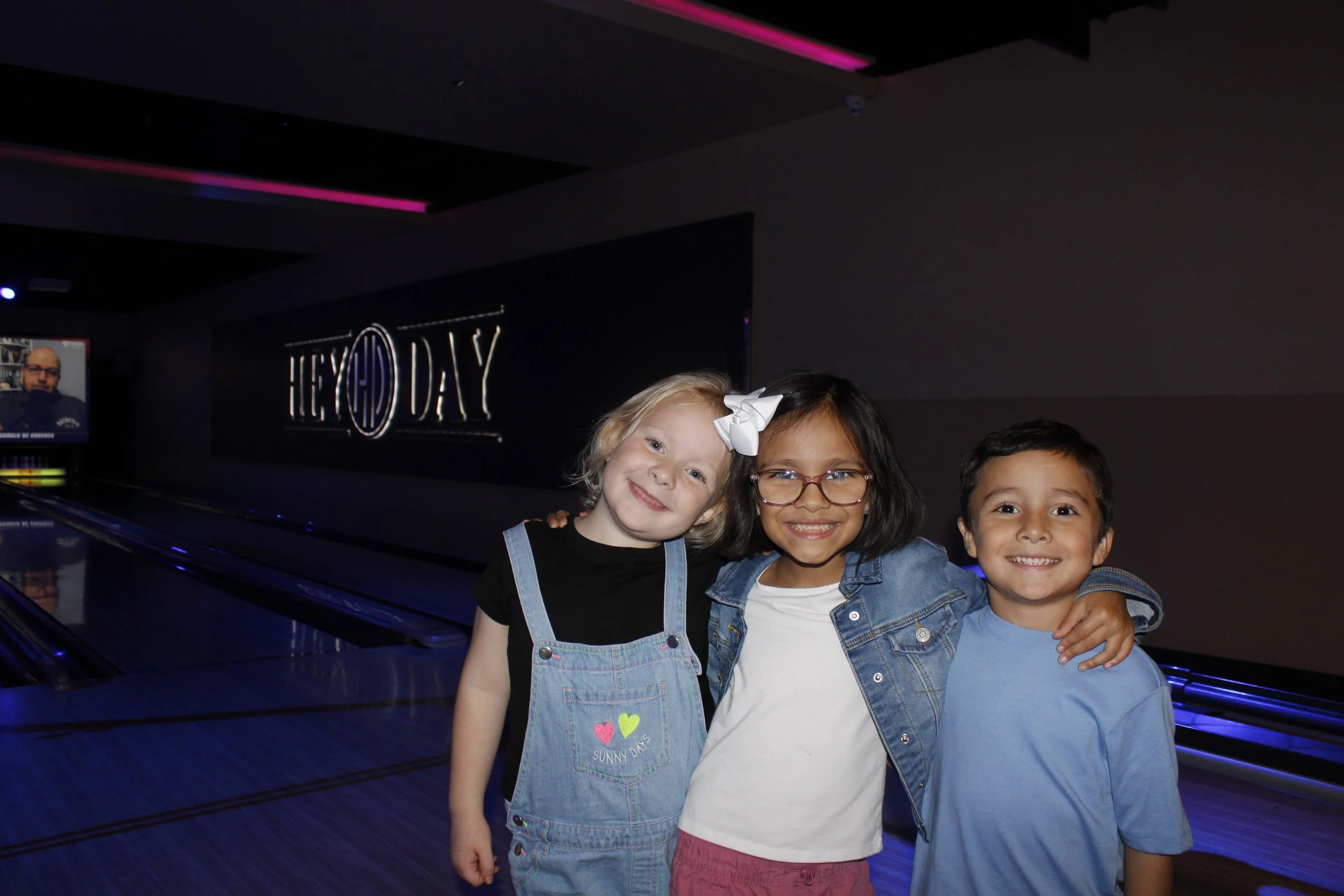 Three children smiling at a bowling alley, standing close together with their arms around each other. The background features a neon sign that says 'HEY DAY' and a bowling lane.