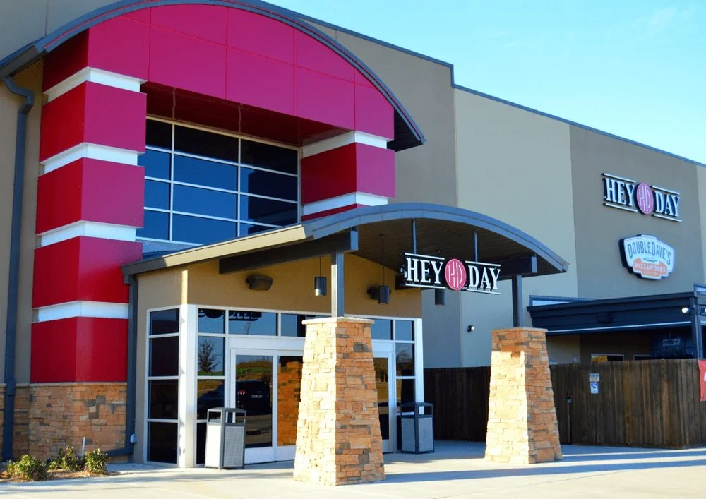 Exterior of a commercial shopping center with entrances to Hey Day and Double Dave's Pizza. The building features red, gray, and beige colors with stone accents and large glass windows.