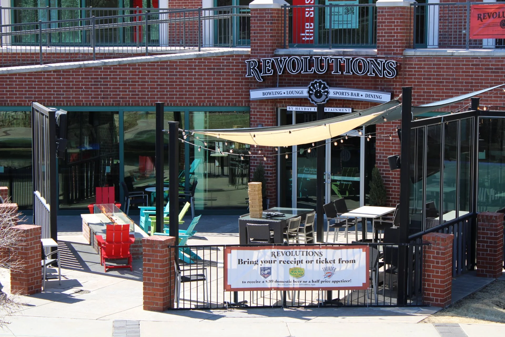 Outdoor patio of Revolutions entertainment venue with colorful chairs, tables, string lights, and a banner that instructs guests to bring receipts for discounted drinks or appetizers. The venue offers bowling, lounge, sports bar, and dining options.