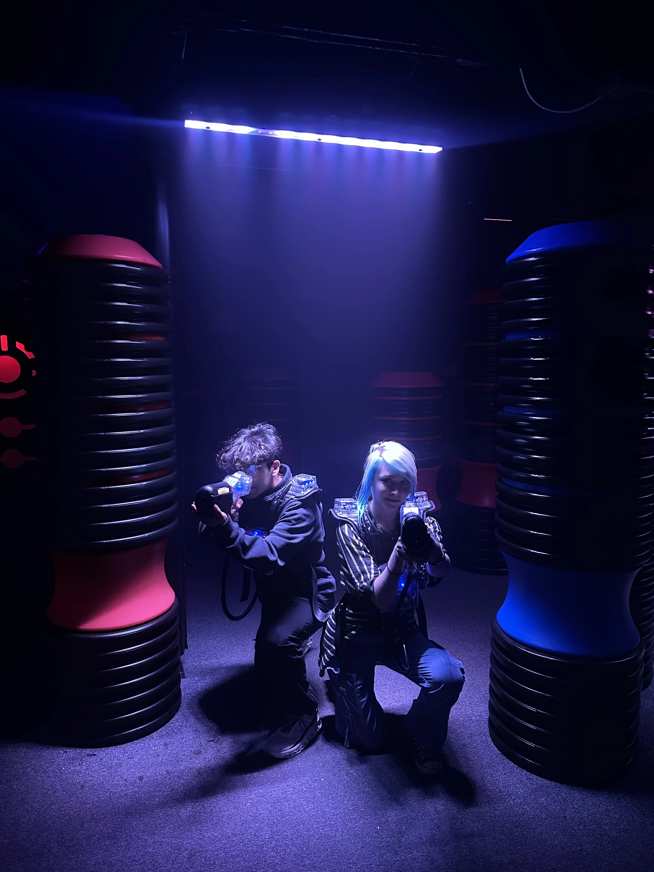 Two people kneeling and holding laser tag guns in a dark room with glowing blue and purple lights and foam barriers.