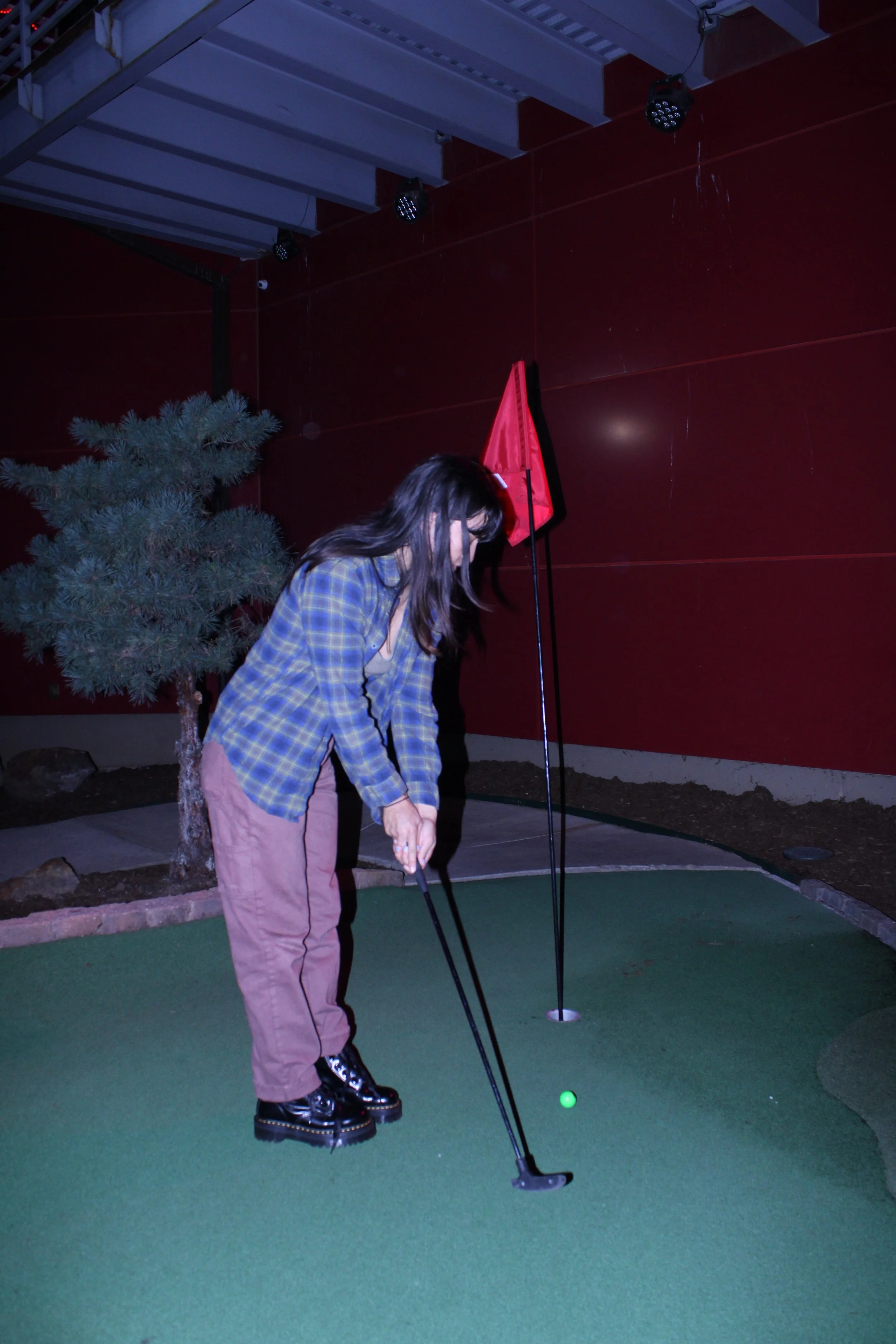 A woman in a plaid shirt and pink pants playing mini golf, preparing to hit a green ball into a hole on an indoor miniature golf course.