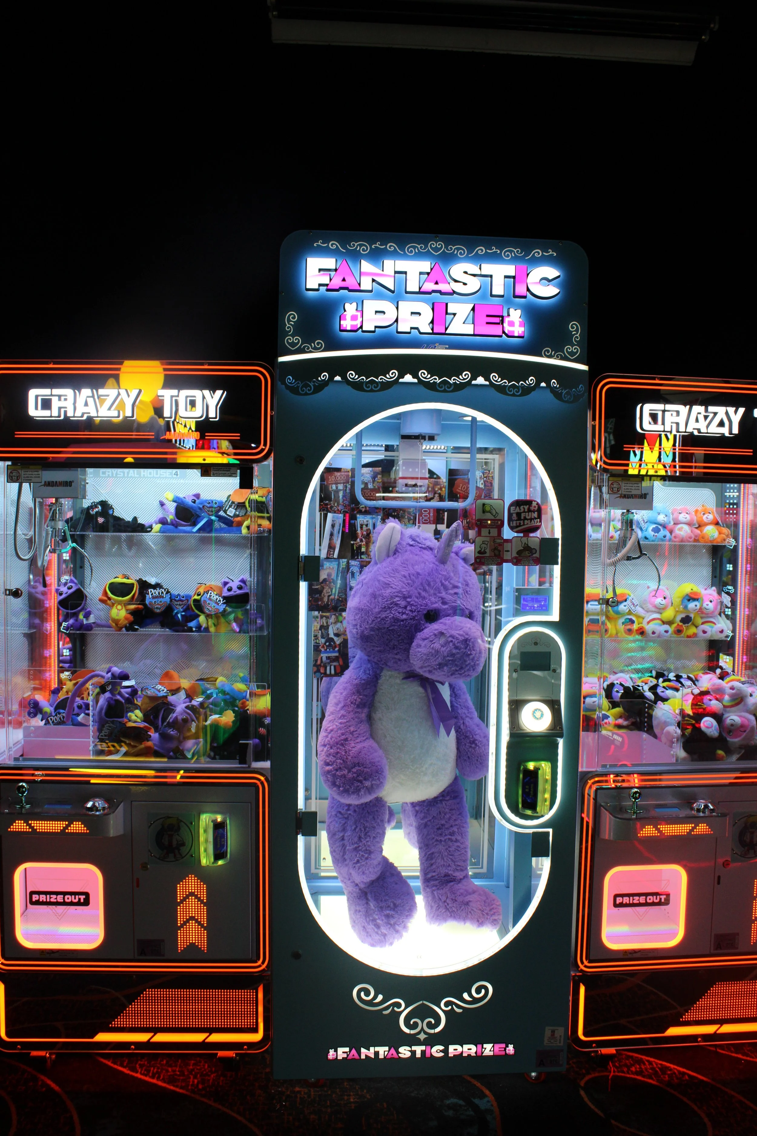A purple plush unicorn toy inside a claw machine labeled 'Fantastic Prize,' with toy-filled machines labeled 'Crazy Toy' on either side at an arcade.