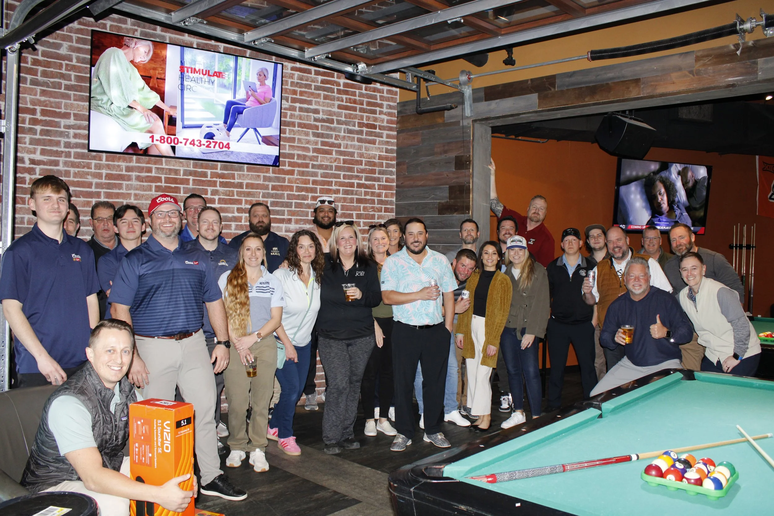 Group of people gathered in a bar or recreational room, some holding drinks, smiling for the camera, with pool table and pool balls in the foreground, and two TVs on brick and wood-paneled walls.