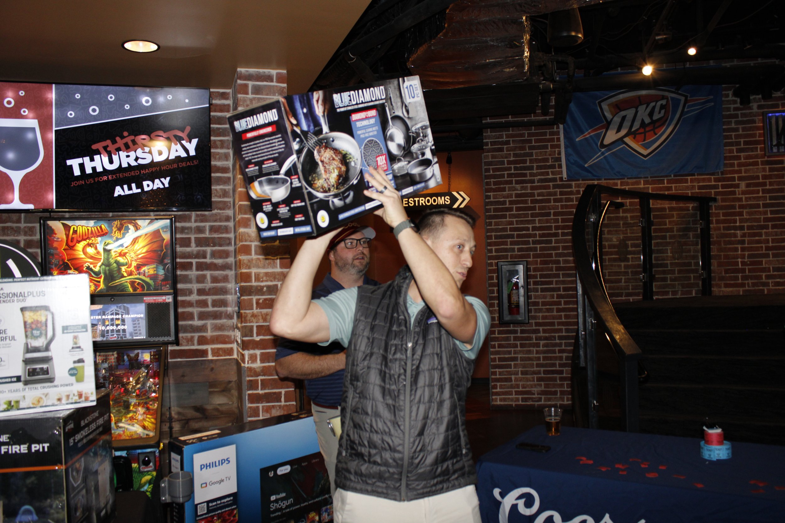 A man in a gray vest and light blue shirt holds up a Blue Diamond almond cookery book in a bar or arcade setting. Behind him, a man with glasses in a striped shirt stands near arcade machines with a Godzilla pinball game. The background shows a brick