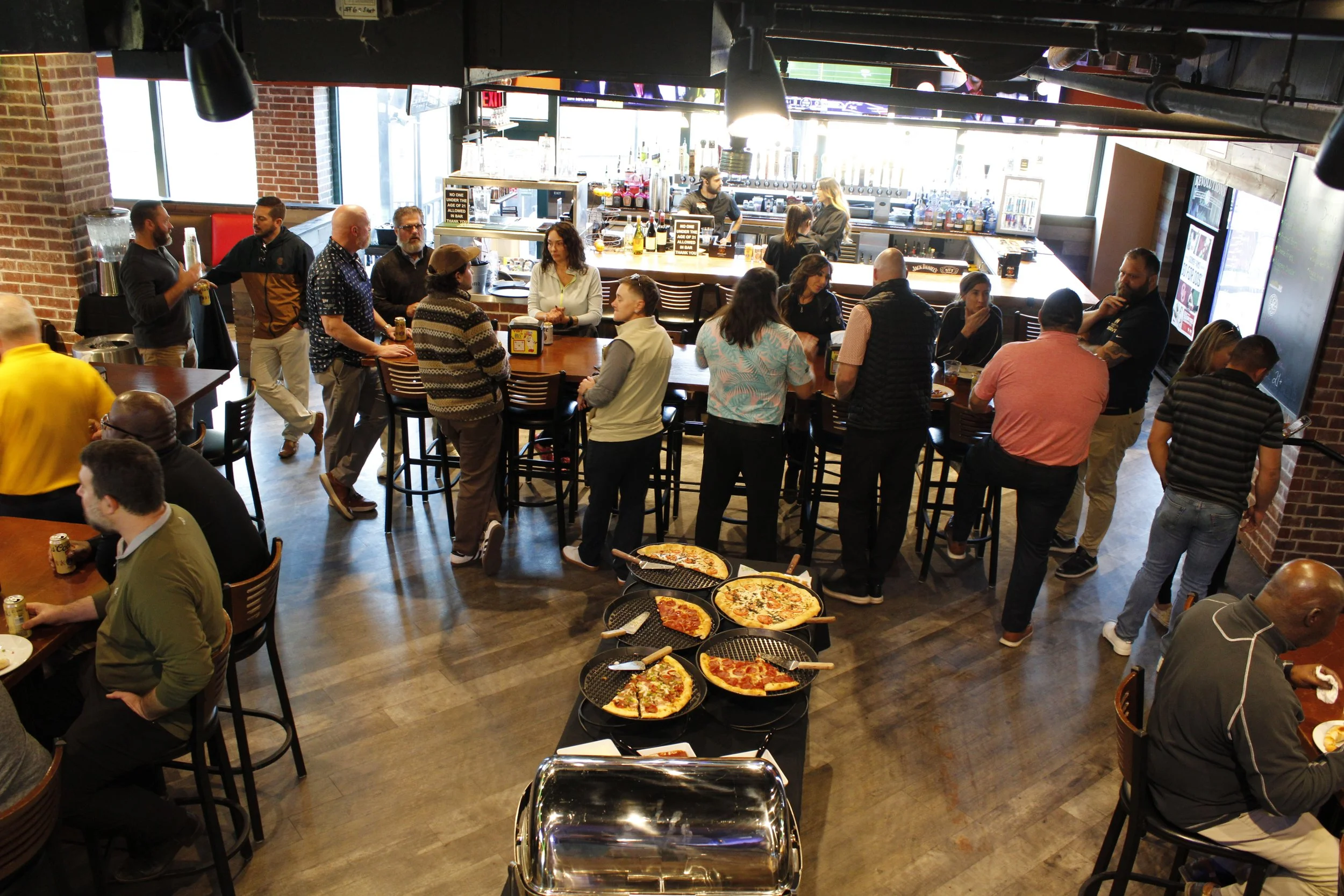 People socializing and eating in a lively restaurant or bar with a pizza station, bar area, and large windows.
