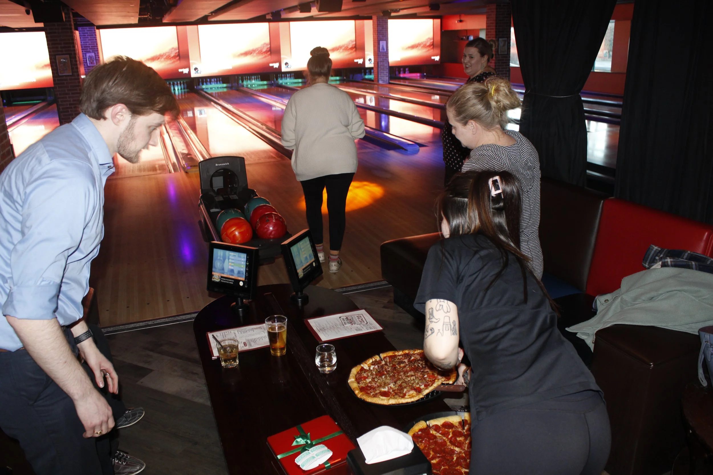 Group of people at a bowling alley, with some preparing to bowl and others enjoying pizza at a table.