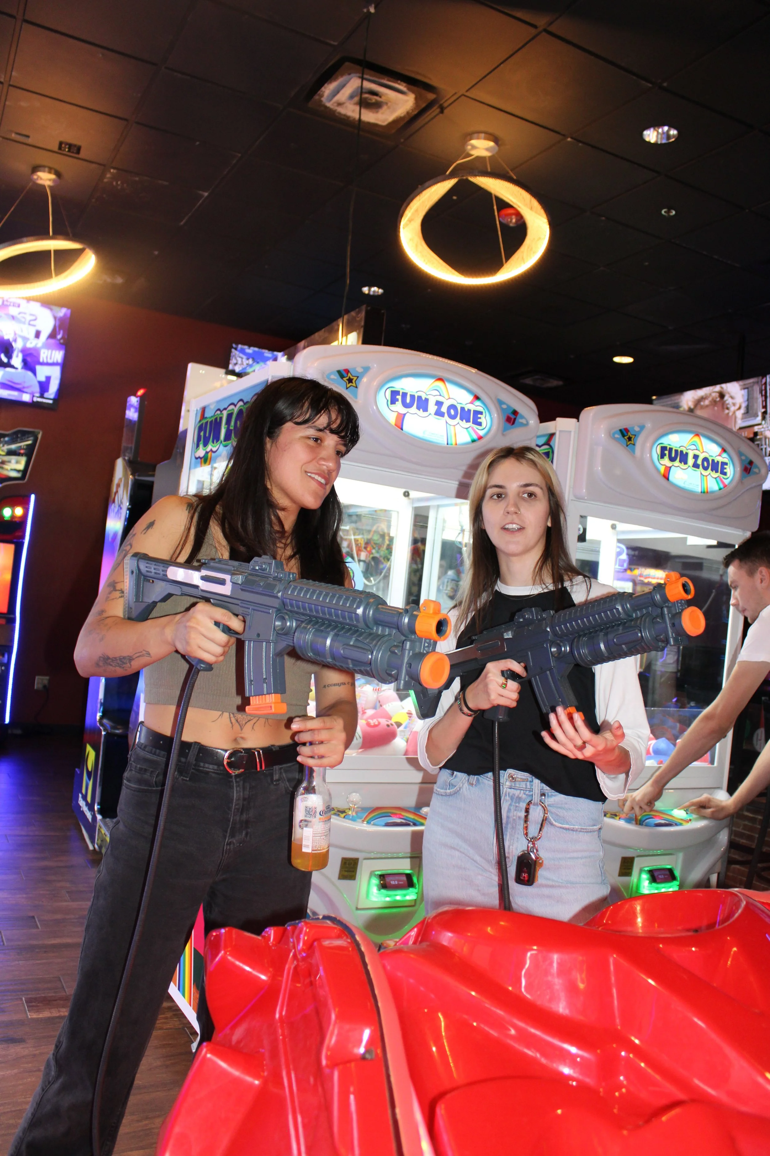 Two women are playing an arcade shooting game in an arcade, each holding toy guns. There are arcade machines and bright lights in the background.