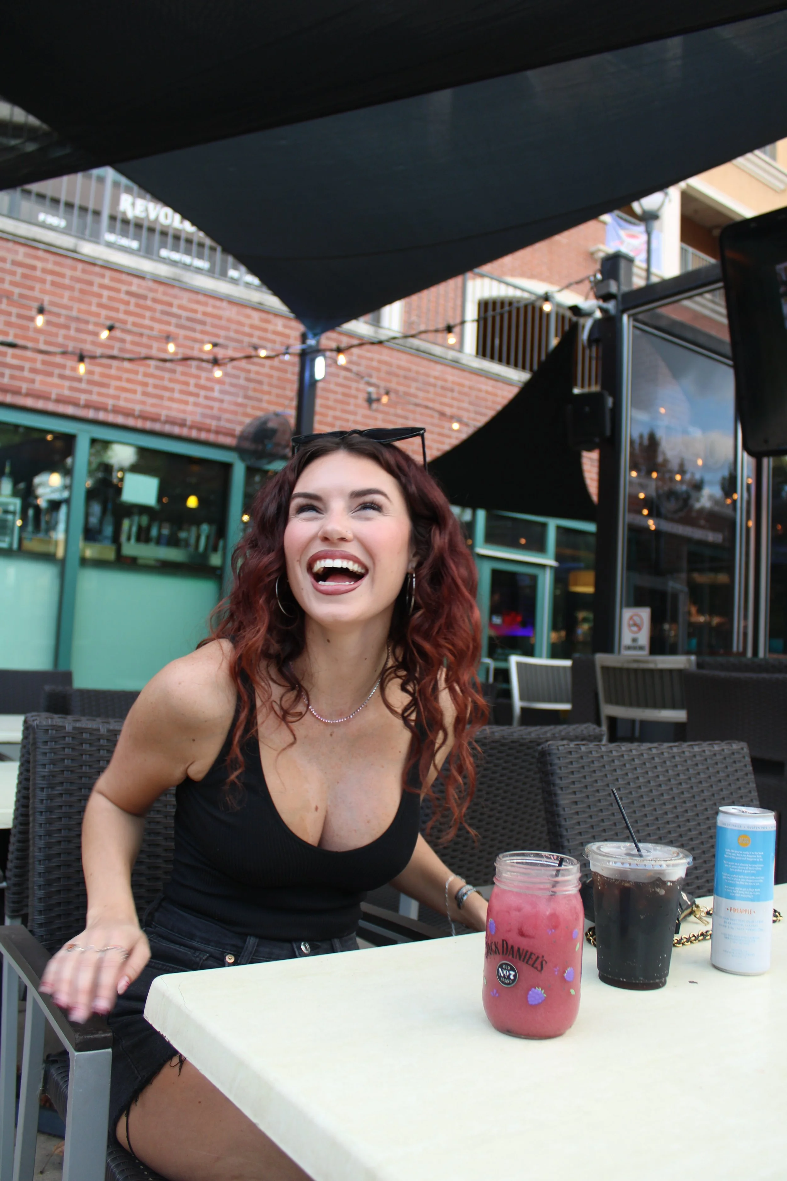 A woman with long curly red hair, wearing a black top and denim shorts, smiling and laughing at an outdoor café. There are drinks in front of her, including a pinkberry drink in a Mason jar and a dark soda, with a can of pineapple-flavored beverage nearby. The background shows a brick building, string lights, and patio furniture.