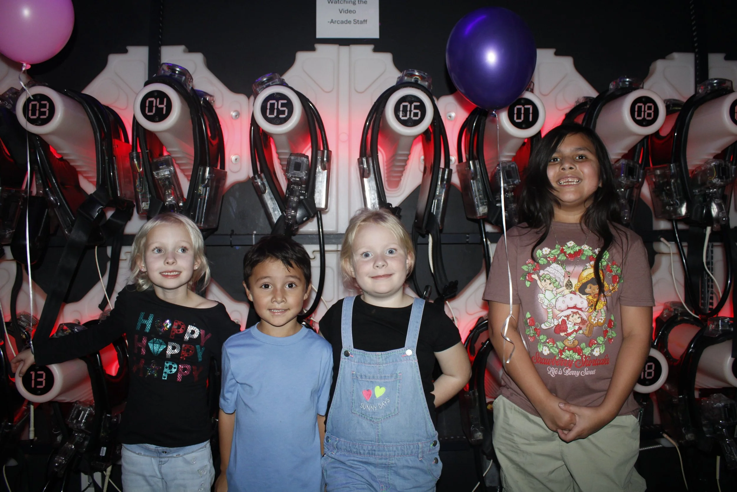 Five children standing in front of a row of arcade racing game machines, smiling.