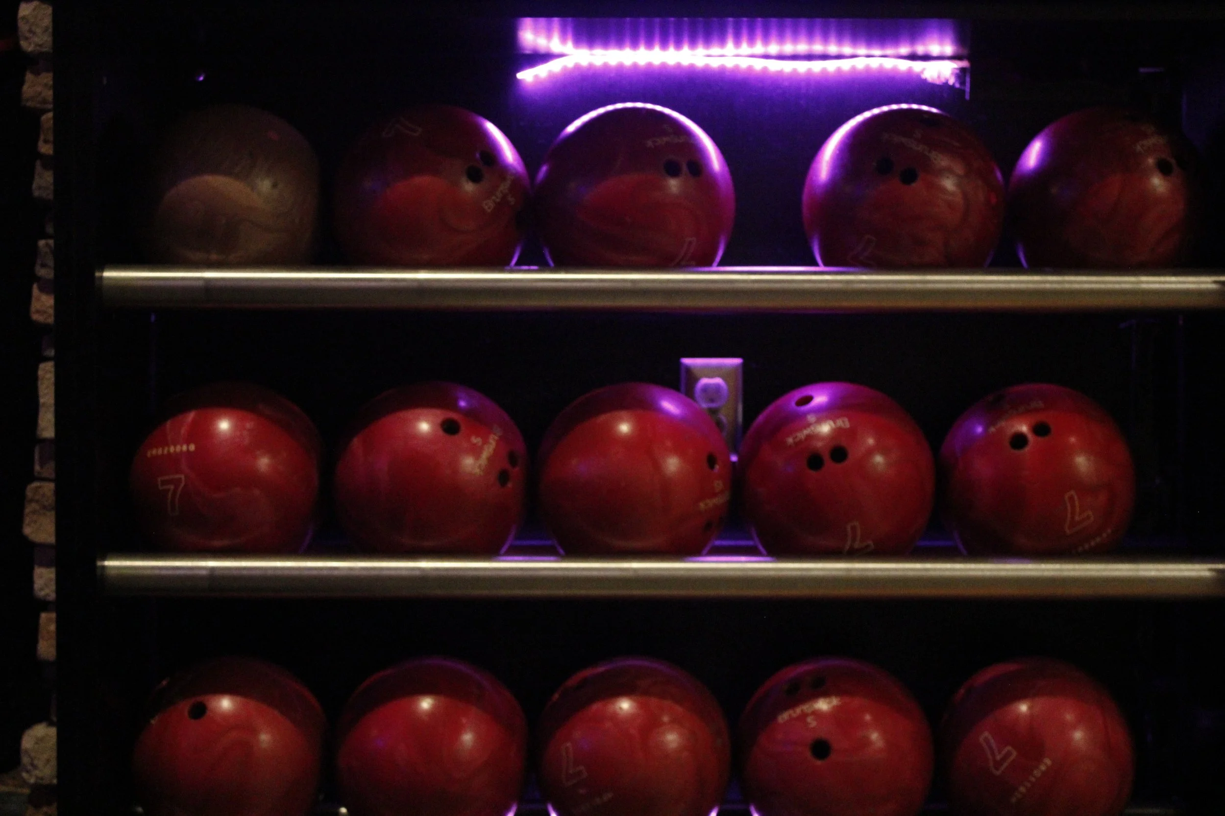 Red bowling balls on a rack illuminated by purple lighting.