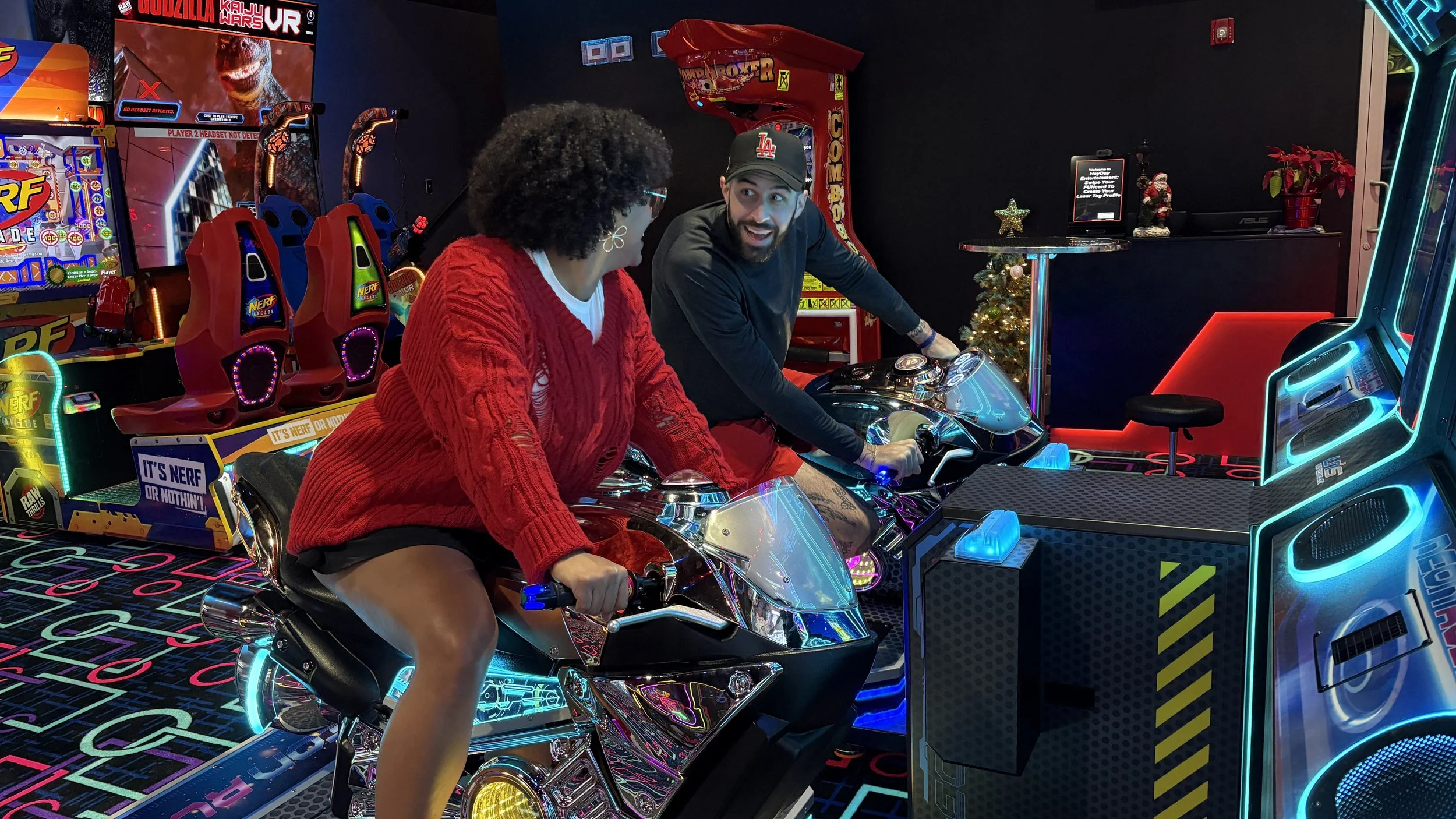 Two people riding arcade motorcycle simulators in an arcade with colorful neon lights and game machines around.