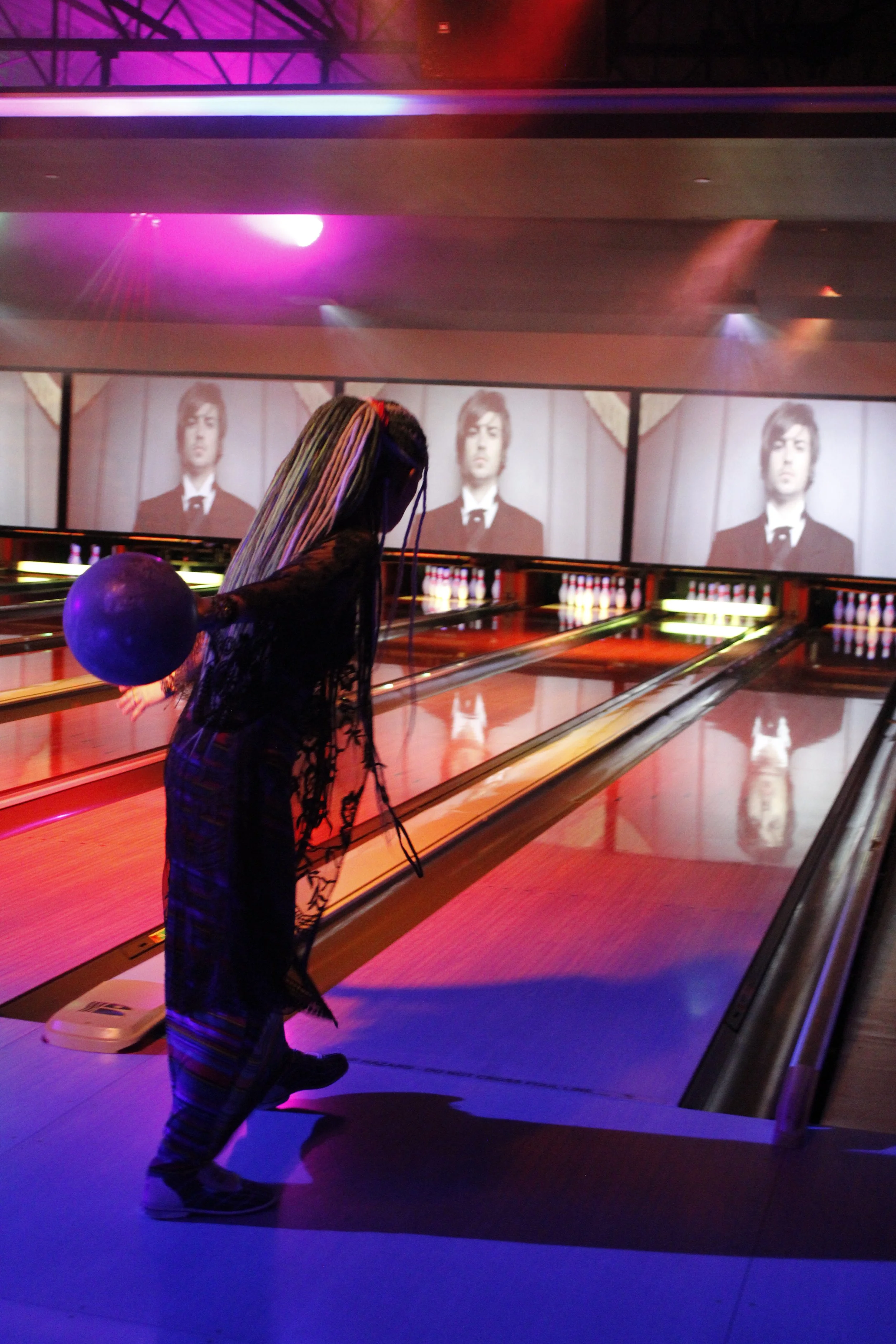 A woman with colorful braided hair and patterned pants holding a blue bowling ball, standing on a bowling alley with colorful lights and a screen displaying a person's face in the background.