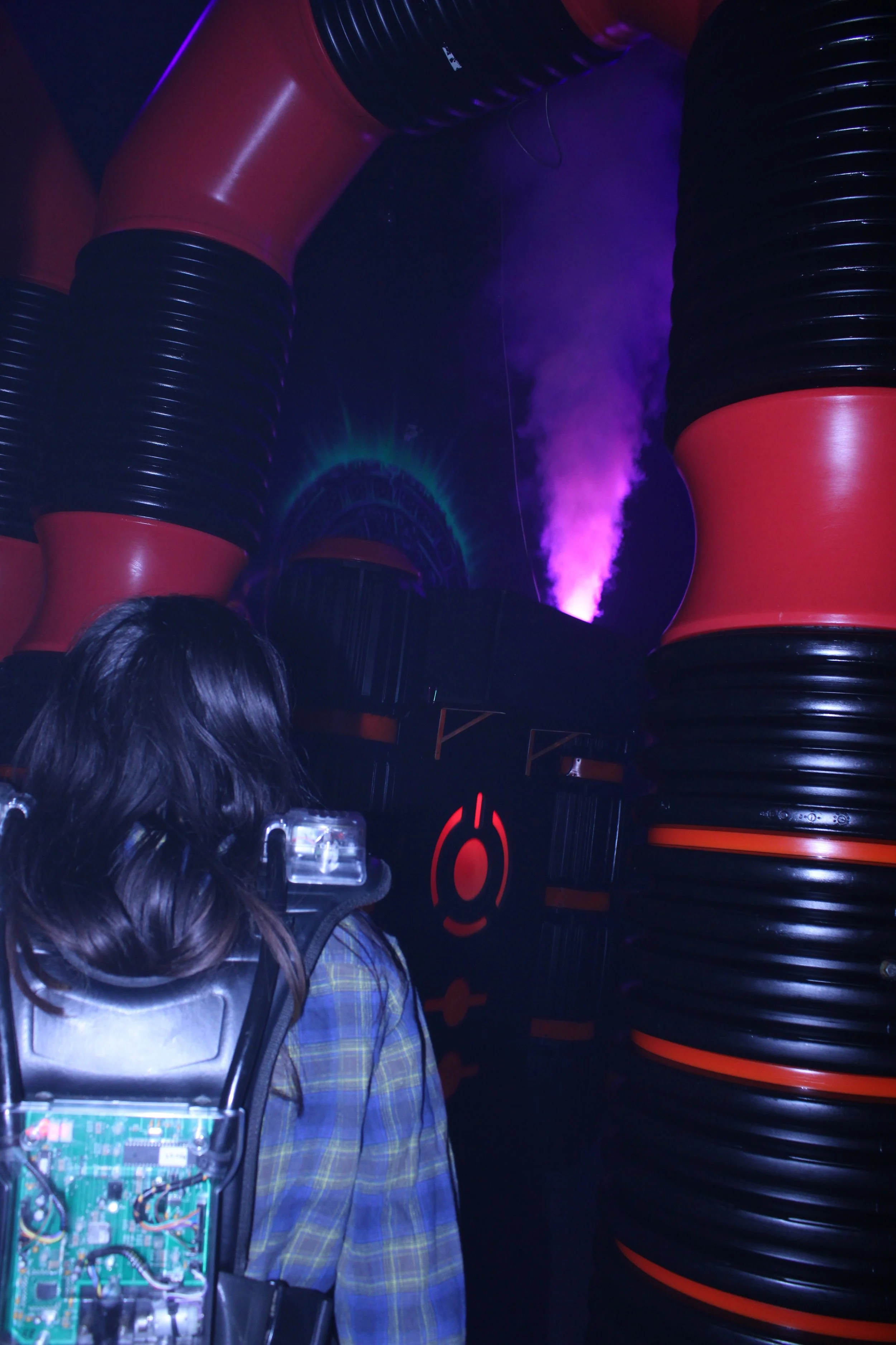 A person with long hair wearing a checkered shirt and carrying a backpack with visible electronics inside, standing in front of large, inflatable tube structures with purple lighting and steam or smoke in the background.