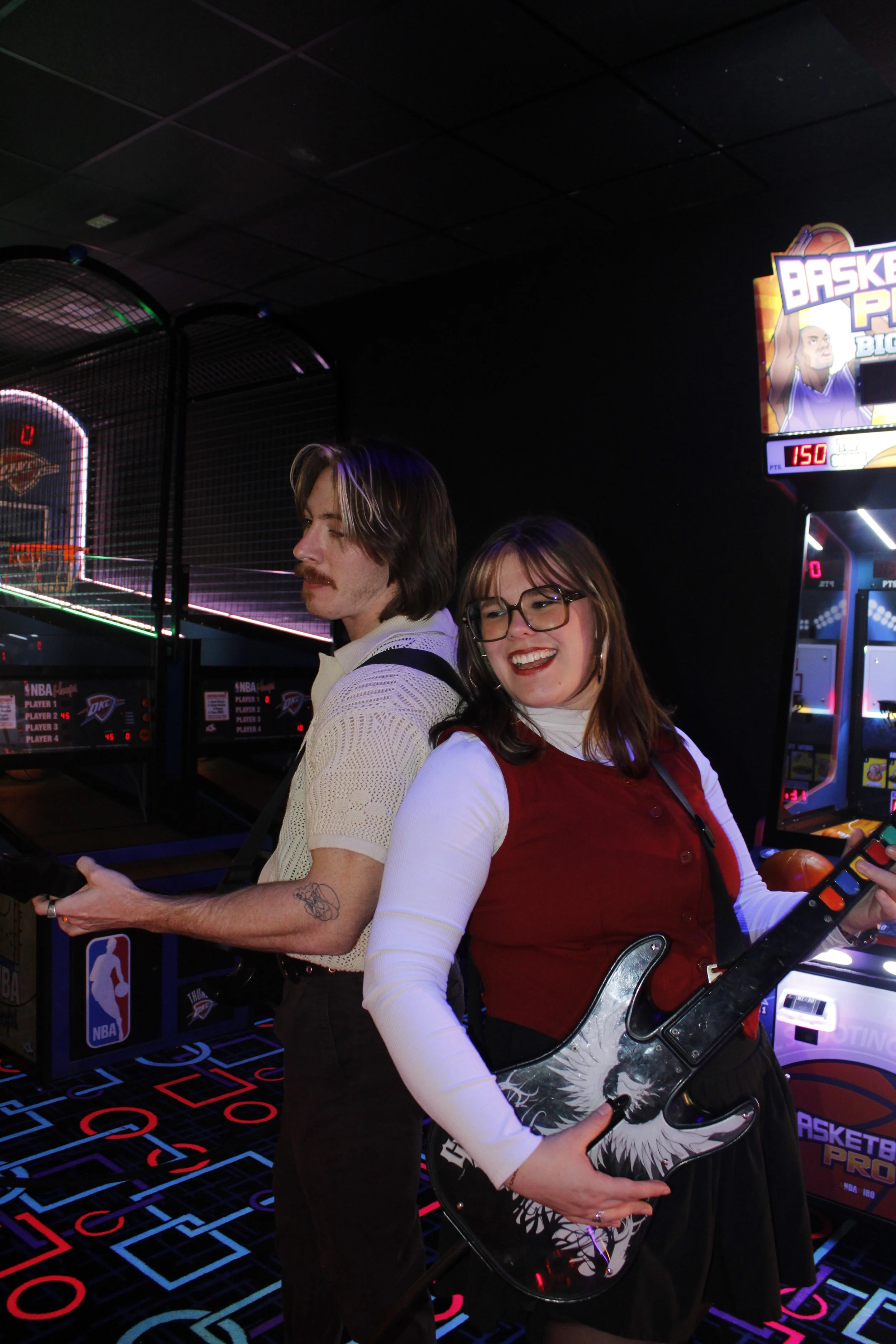Two people, a man with brown hair and a mustache wearing a cream-colored sweater and a woman with glasses and long brown hair in a black dress, standing in an arcade next to ping pong arcade games and holding a guitar-shaped arcade controller, with basketball arcade machines in the background.