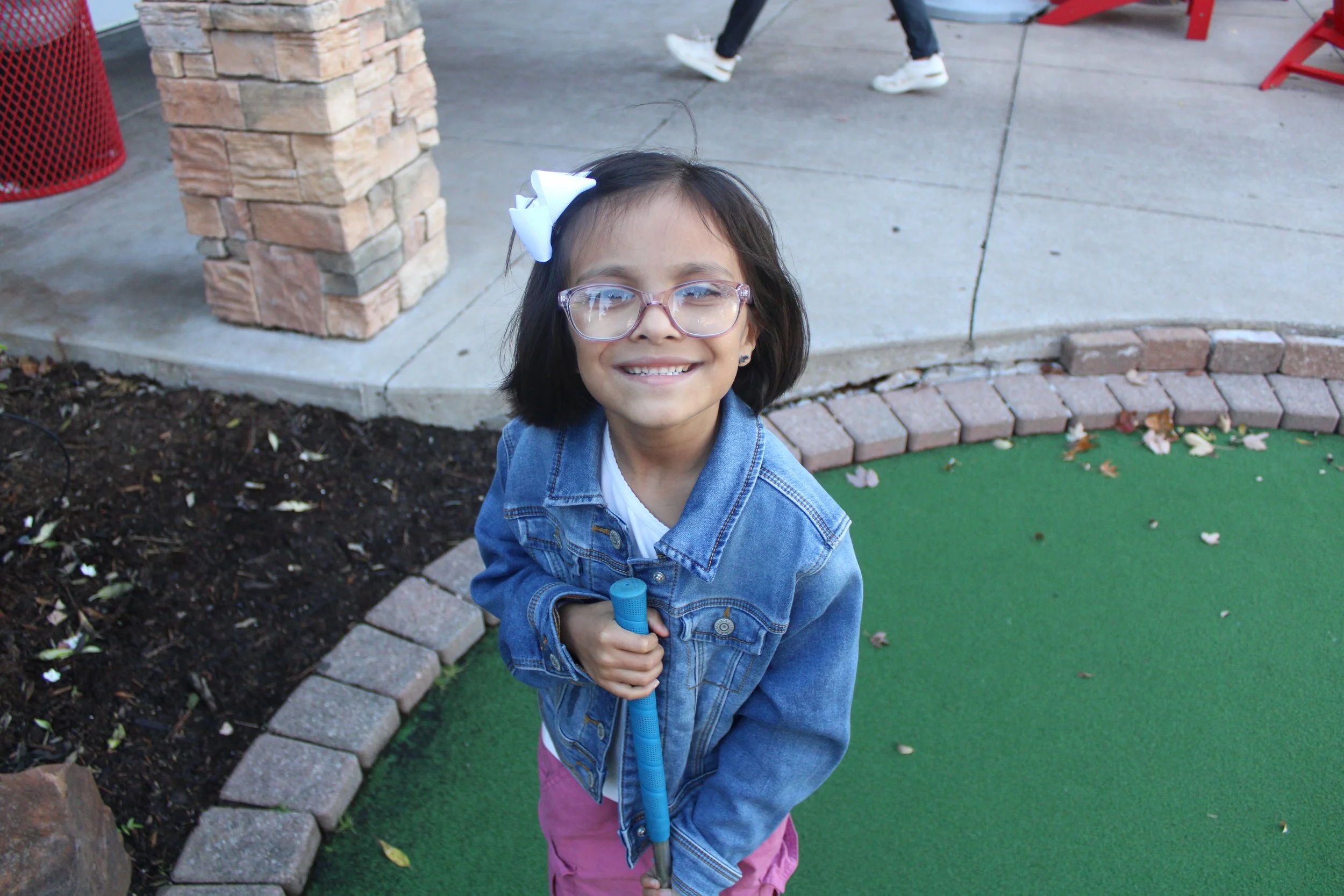 A young girl with glasses smiling at the camera, wearing a denim jacket and holding a blue toy, standing outside near a brick-paved area with a green artificial grass and a flower bed.