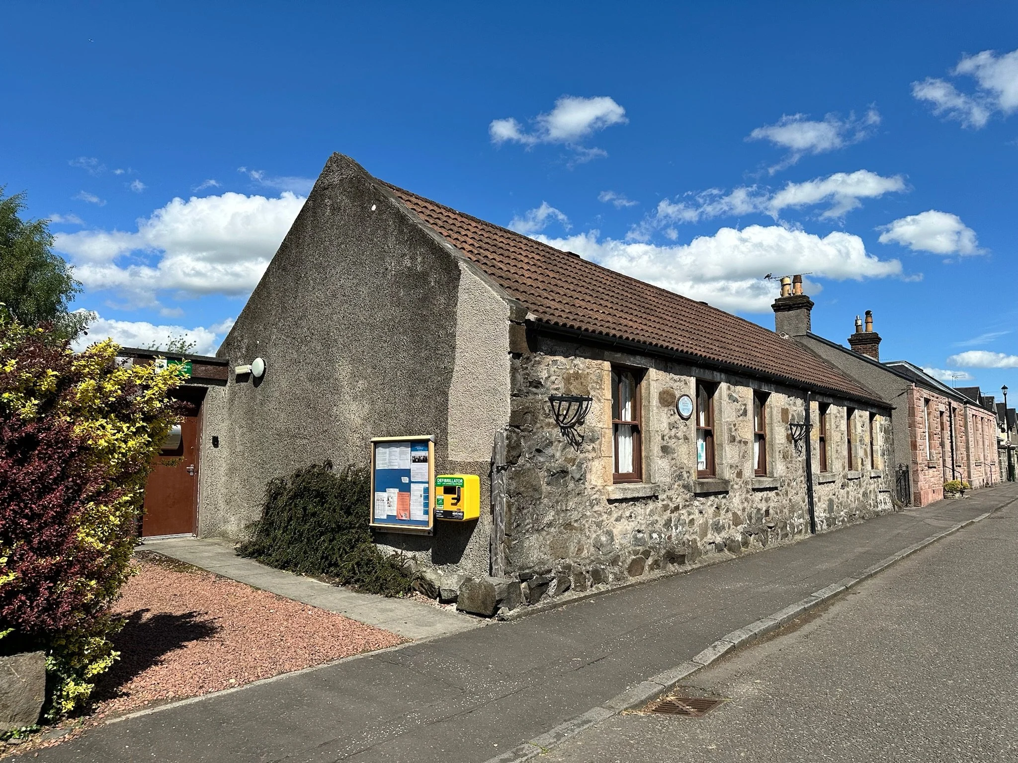 A stone building with a red tile roof and chimney pipes under a partly cloudy blue sky. There are bushes and informational signs near the sidewalk.