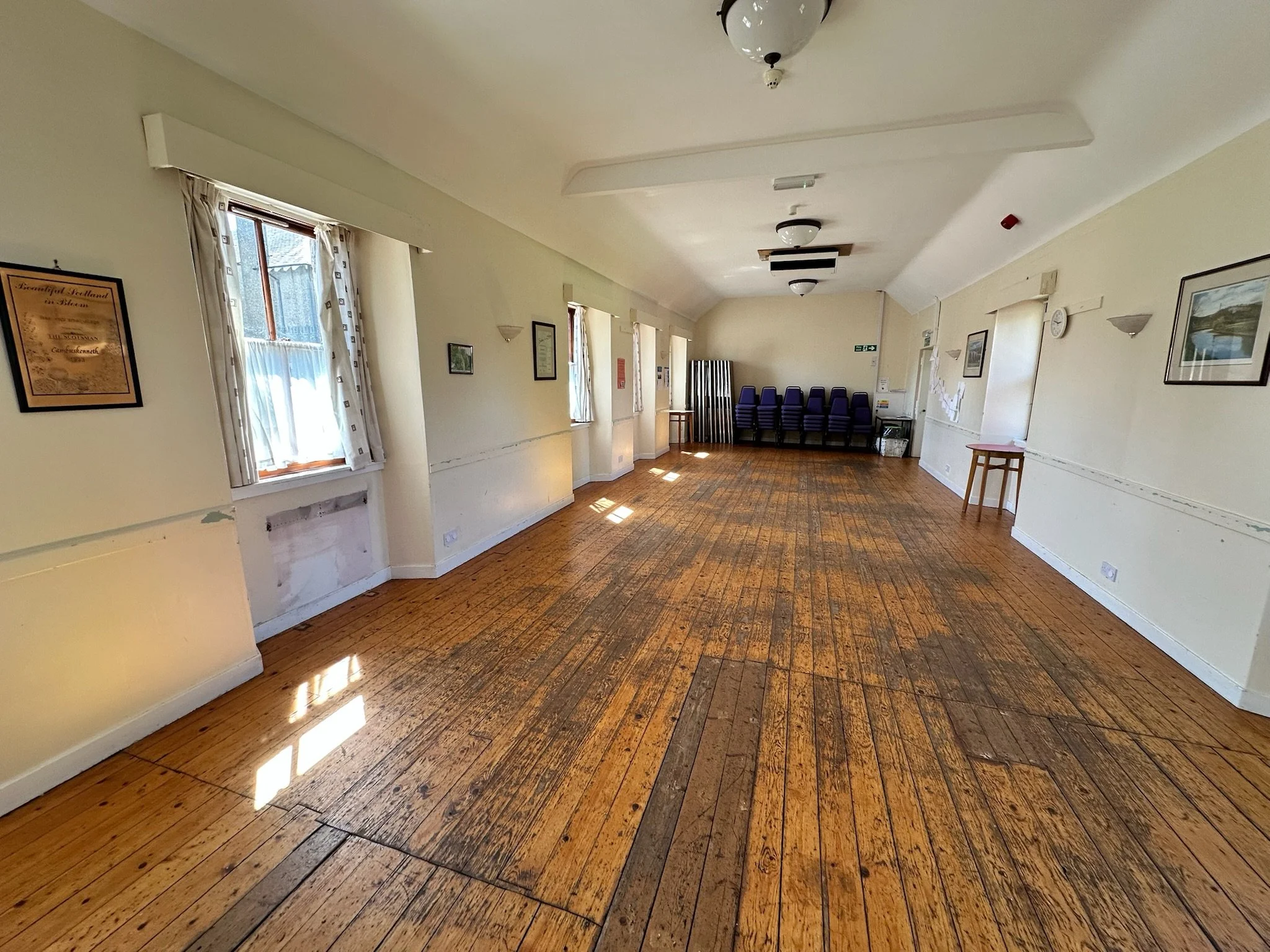 An empty room with wooden floor, white walls, windows with curtains, chairs stacked in one corner, and framed pictures on the walls.