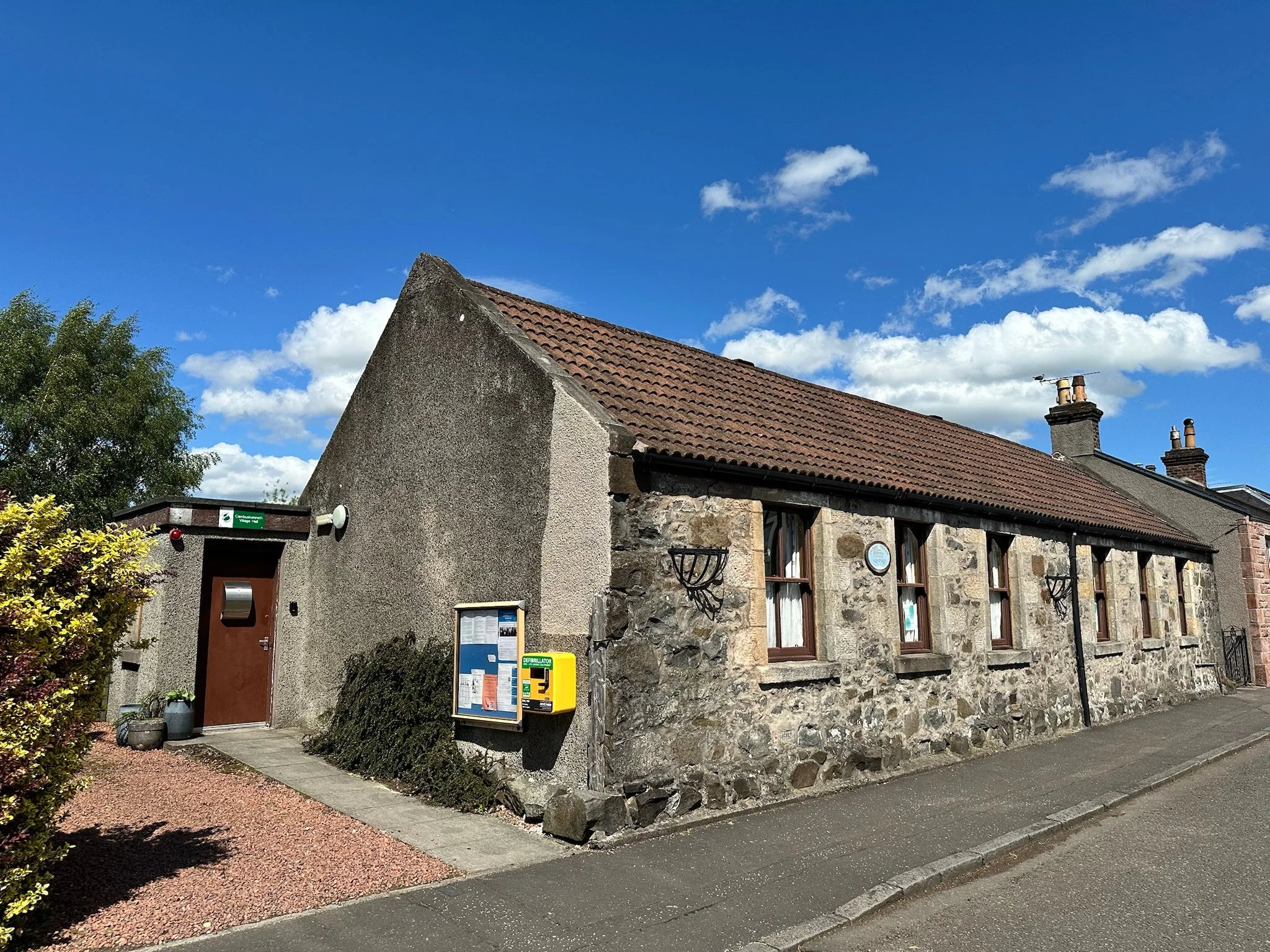 A stone building with a weathered exterior and a terracotta tile roof under a bright blue sky with scattered clouds. There is a greenery bush on the left and a sidewalk in front of the building. A street sign and some notices are visible near a door 