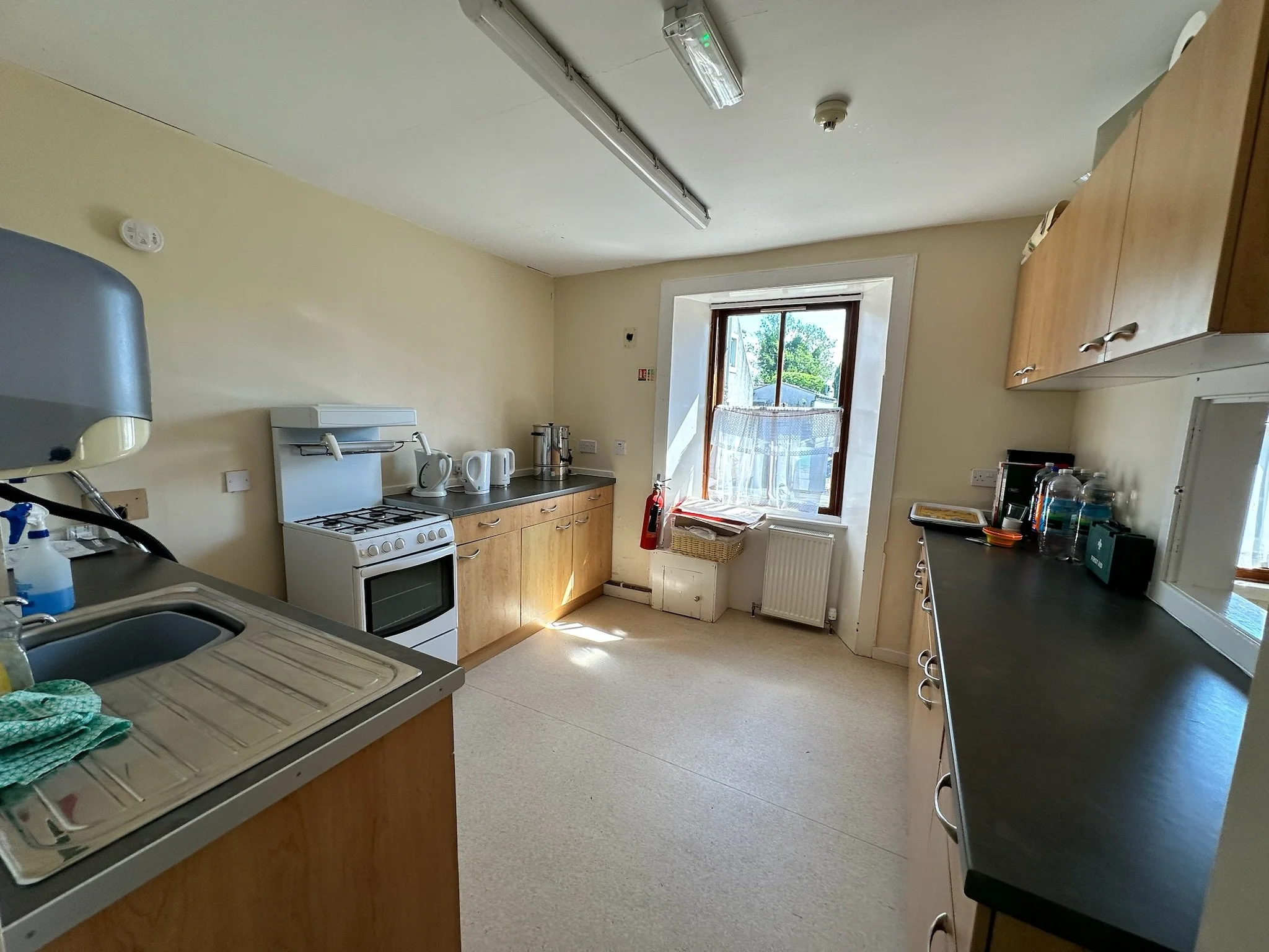 A small kitchen with beige walls and a window letting in natural light. The kitchen has a countertop with various appliances including a microwave, kettles, and coffee makers. There is a stove with a small oven, cabinets above, and a sink counter wit