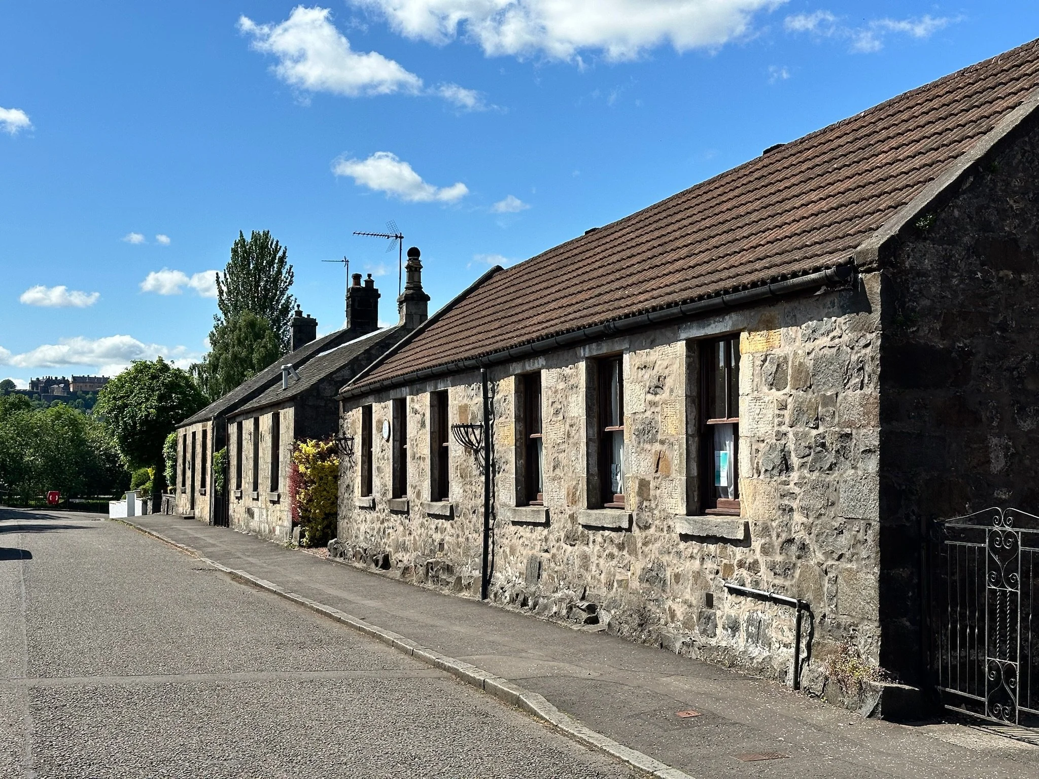A row of old stone buildings with red tiled roofs, chimney stacks, and chimneys along the street under a bright blue sky with scattered clouds.