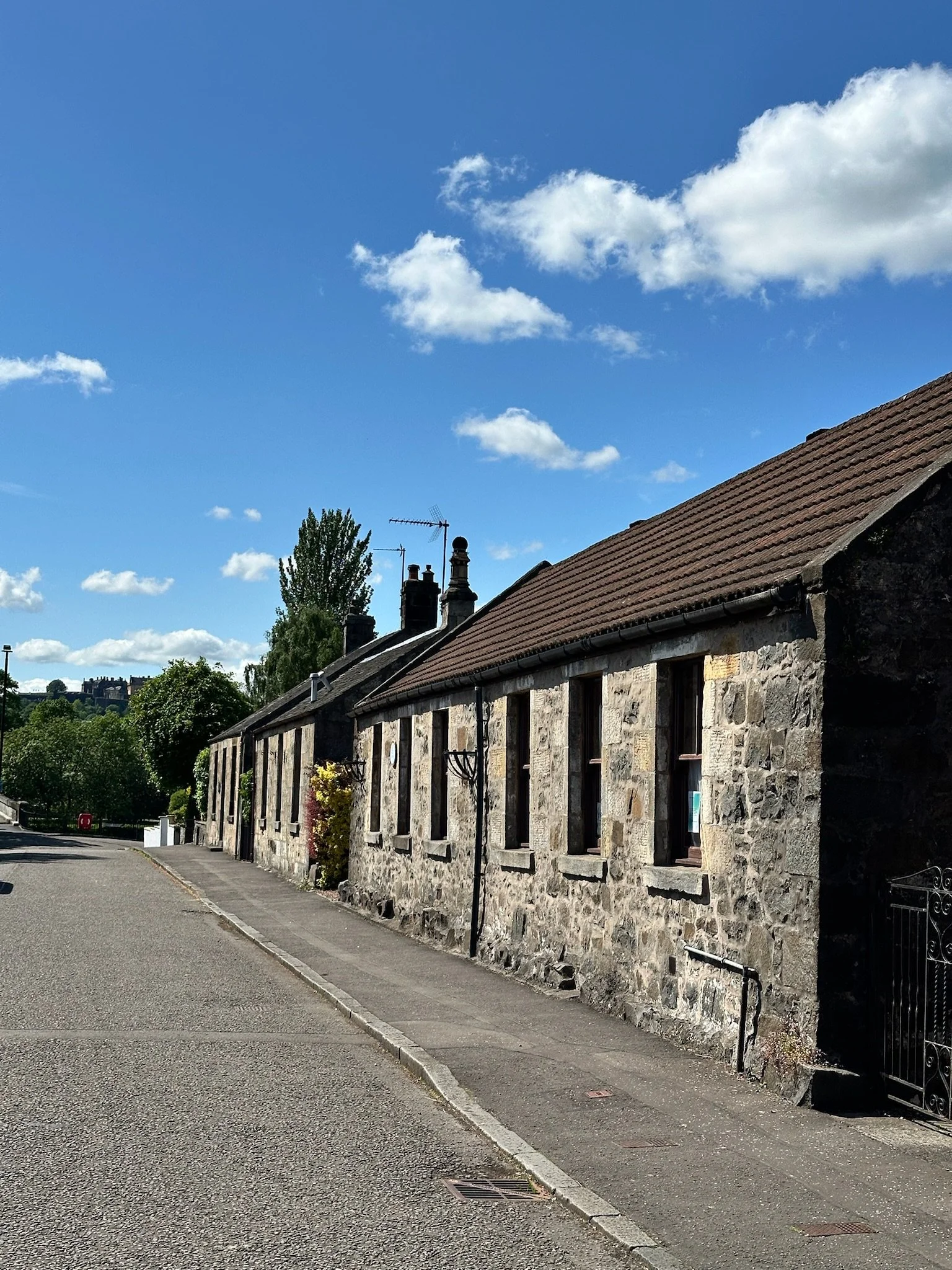 A row of stone houses with tiled roofs along a sidewalk on a sunny day, with green trees and a blue sky with scattered clouds in the background.