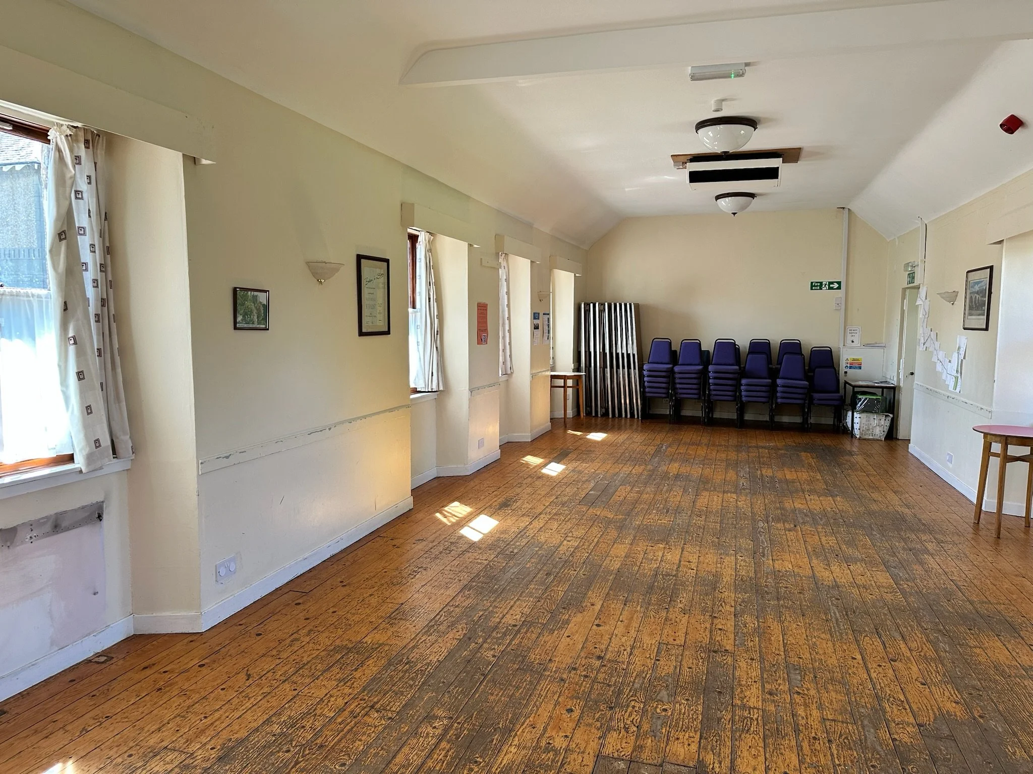 An empty room with hardwood floors, cream-colored walls, and a row of stacked purple chairs along the right wall. There are three windows with white curtains on the left wall, and framed pictures hanging on the walls. The ceiling has two dome-shaped 