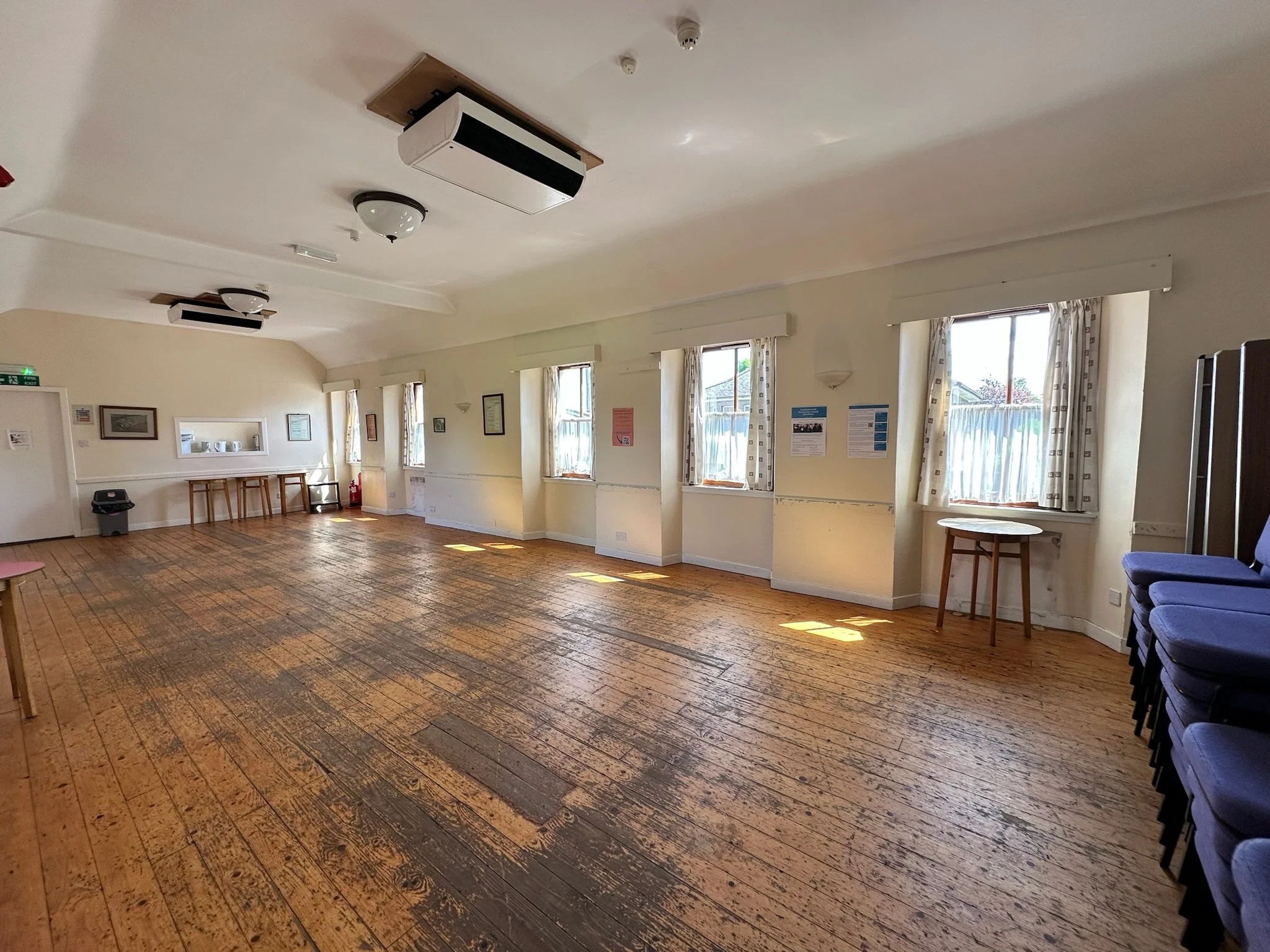 An empty room with four windows on the right wall, cream-colored walls, hardwood flooring, and ceiling lights. There are blue chairs stacked on the far right side and a small table near the window.