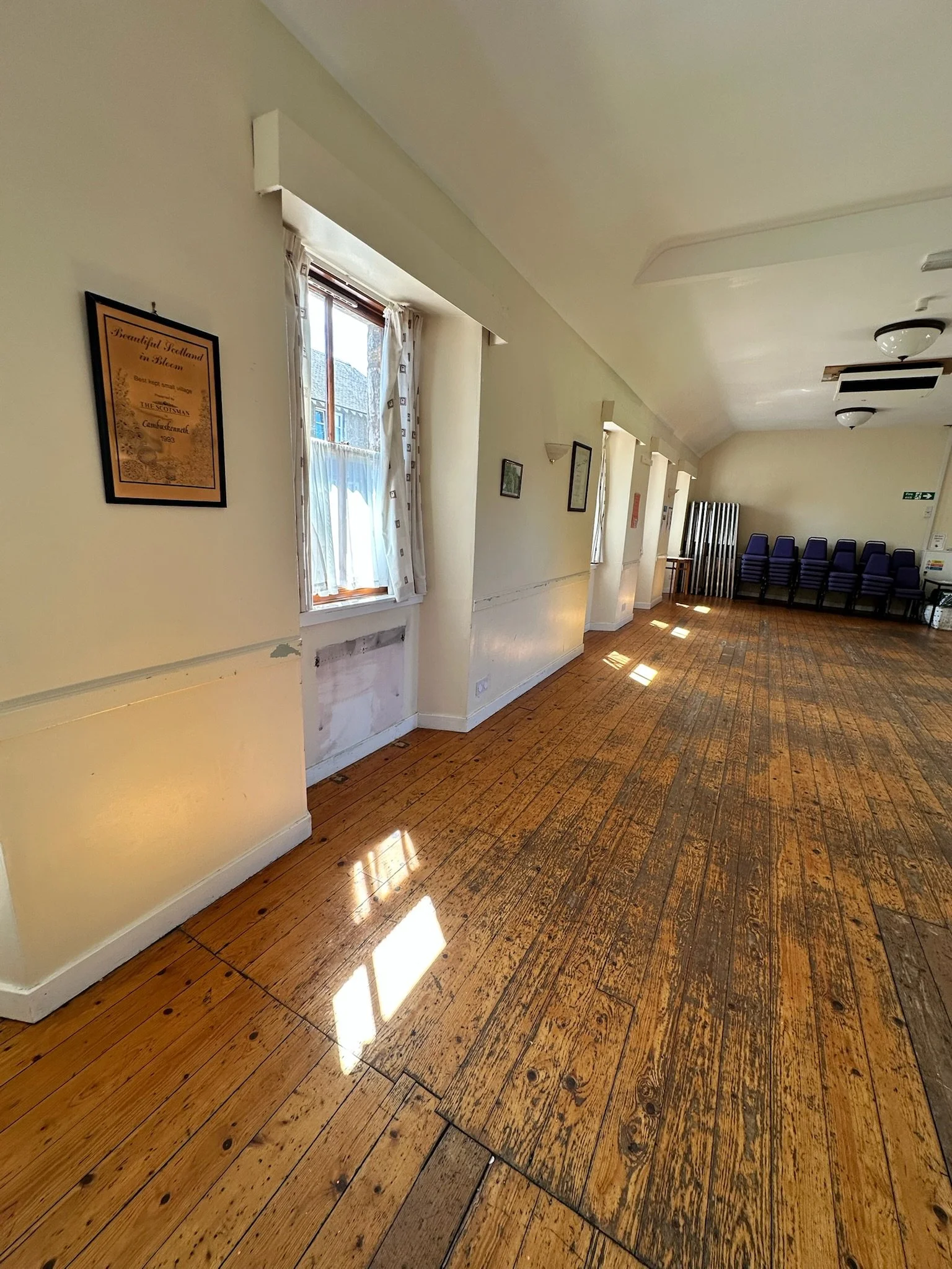 A hallway with wooden floors, cream-colored walls, and a row of stacked purple chairs at the far end. The hallway is lit by natural light coming through a window with white curtains.