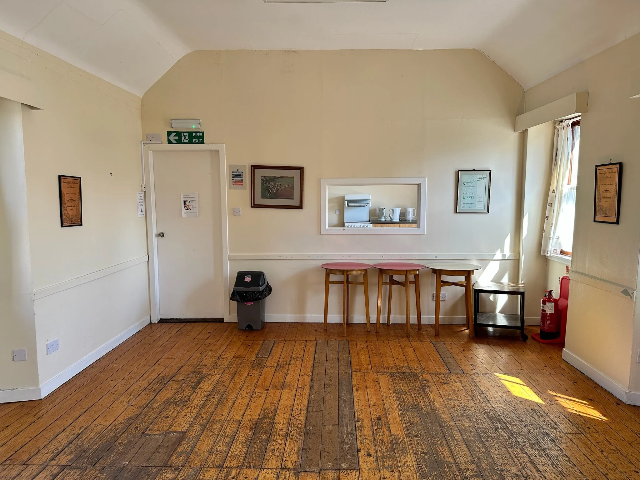The image shows a room with wooden flooring, off-white walls, and a white ceiling with some cracks. There is a small pass-through window in the wall, with a small kitchen area visible through it. The kitchen has a stainless steel dishwasher, electric