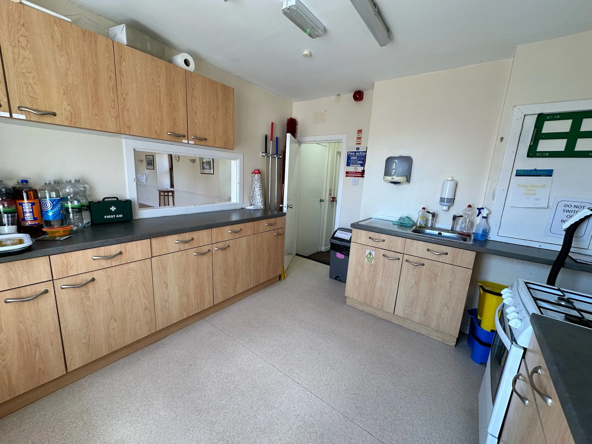 A room with light wood cabinets on the left, a counter with bottles, a first aid kit, and a window with a pass-through opening. There are cleaning supplies, a sink, and a wall-mounted paper towel dispenser. Various cleaning tools and equipment are vi